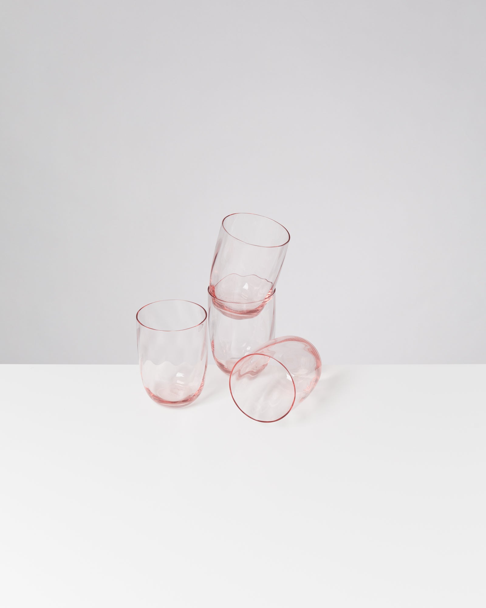 Drei rosa Gläser aus Glas stehen auf einem weißen Tisch, eines leicht gekippt.