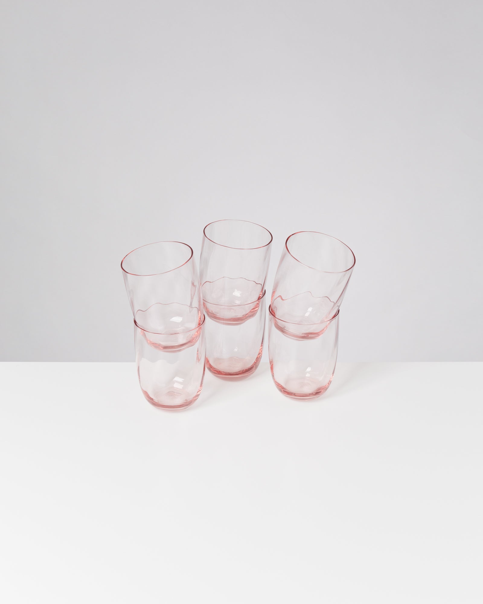 Vier rosafarbene Gläser stehen auf einem weißen Tisch vor einem grauen Hintergrund.