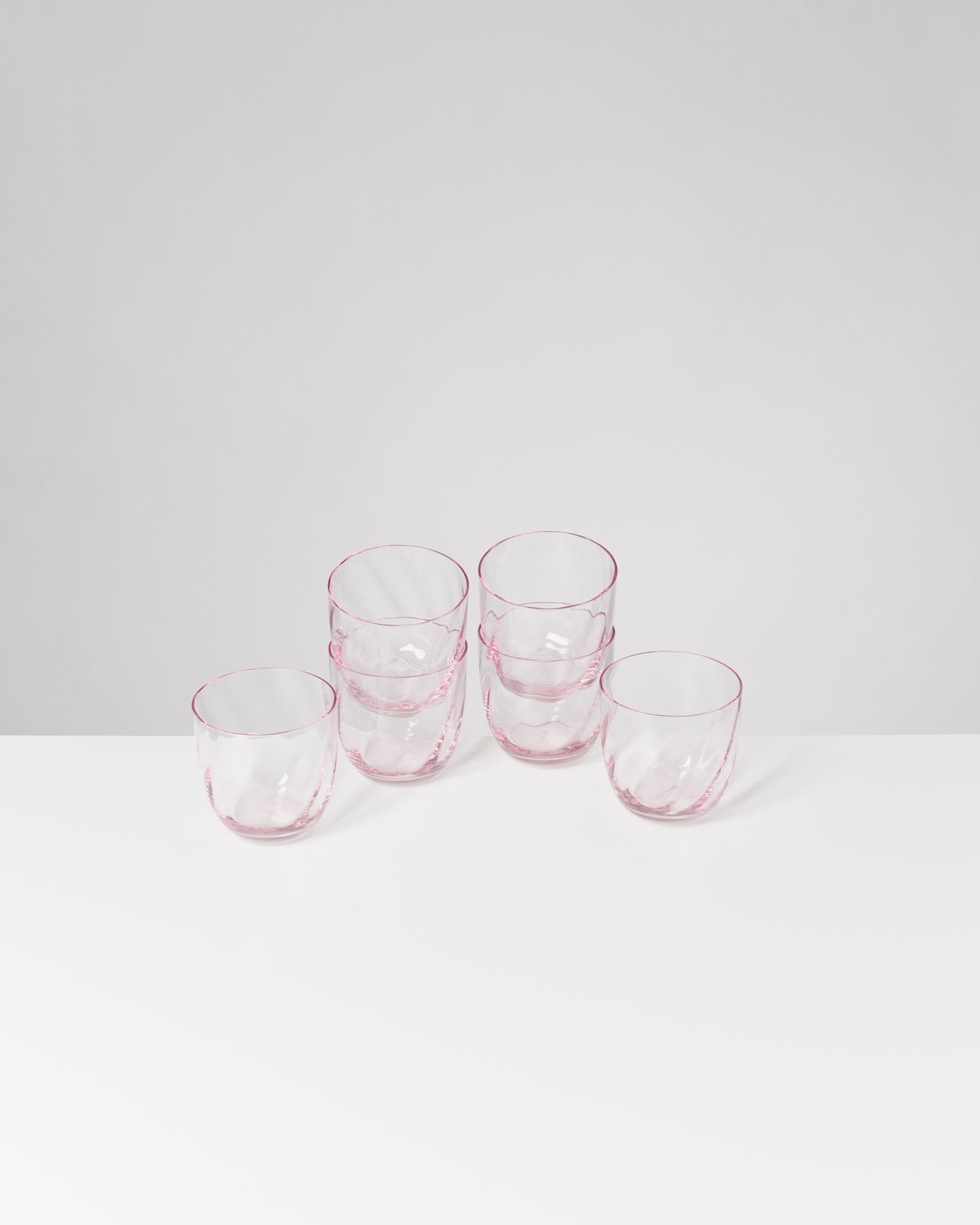 Sechs rosafarben umrandete Gläser auf einem weißen Tisch vor einem grauen Hintergrund.