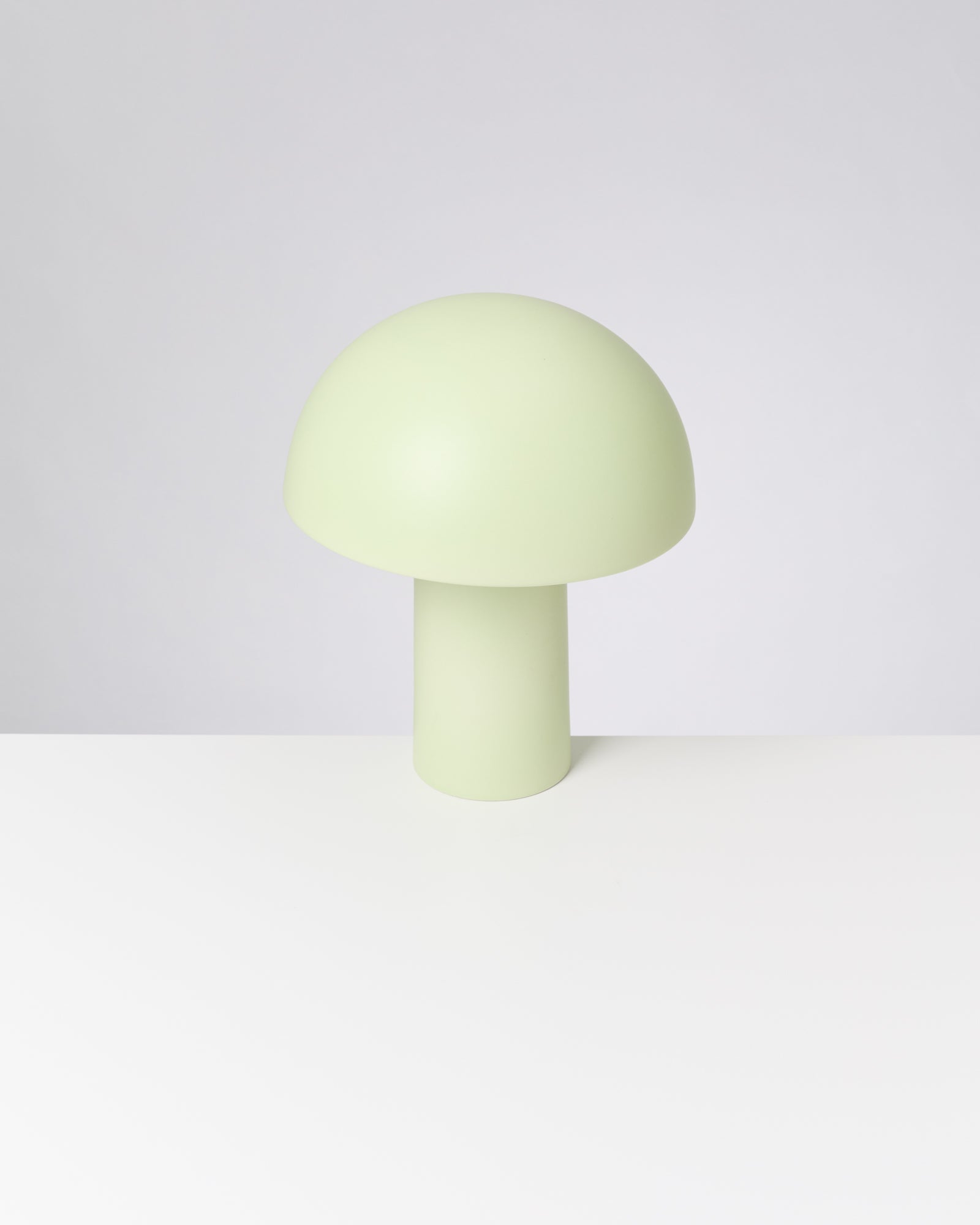 Hellgrüne, pilzförmige Tischlampe auf einem weißen Tisch vor grauem Hintergrund.