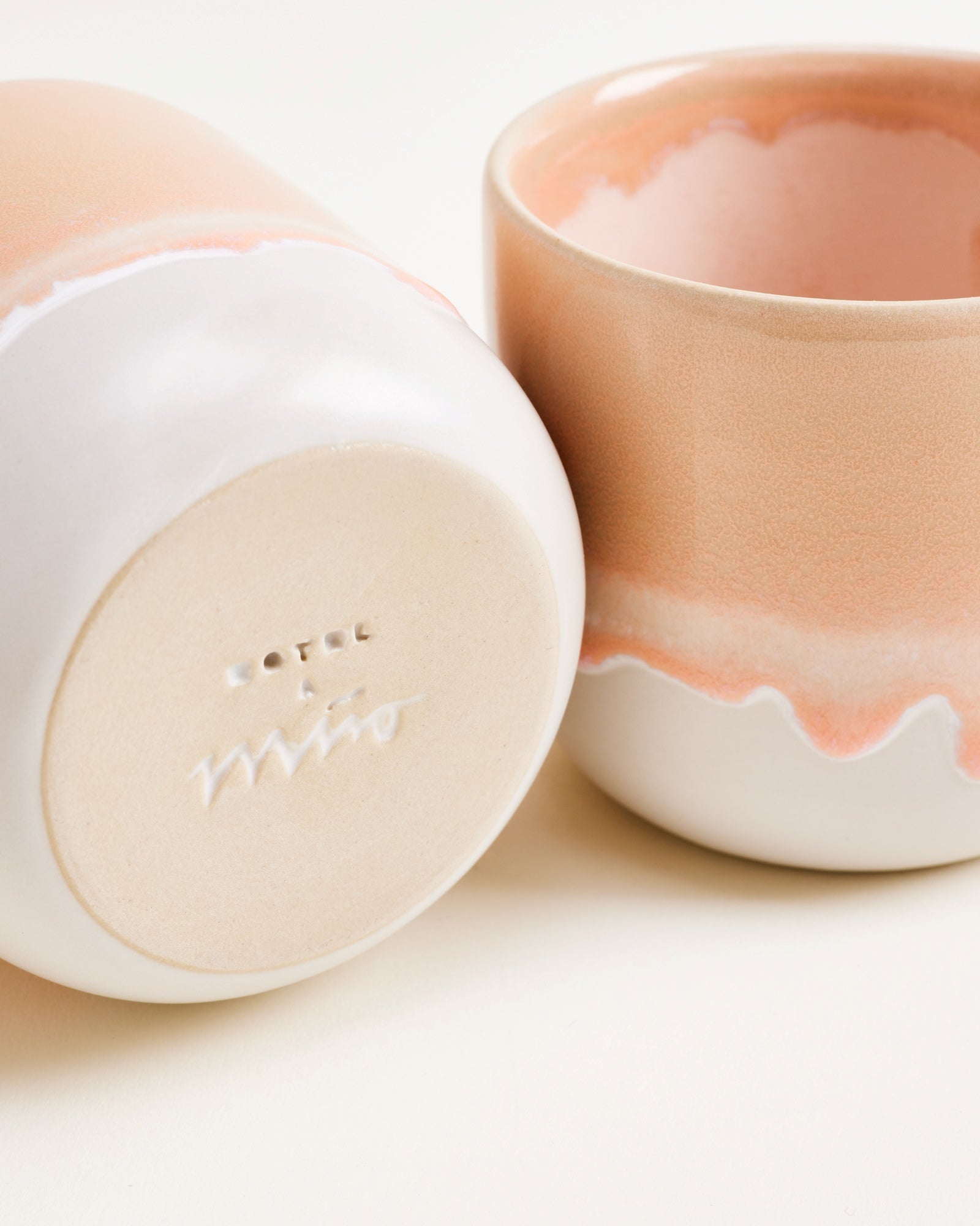 Zwei Keramiktassen in Pastellfarben, eine steht aufrecht, die andere liegt auf der Seite und zeigt den Bodenstempel.