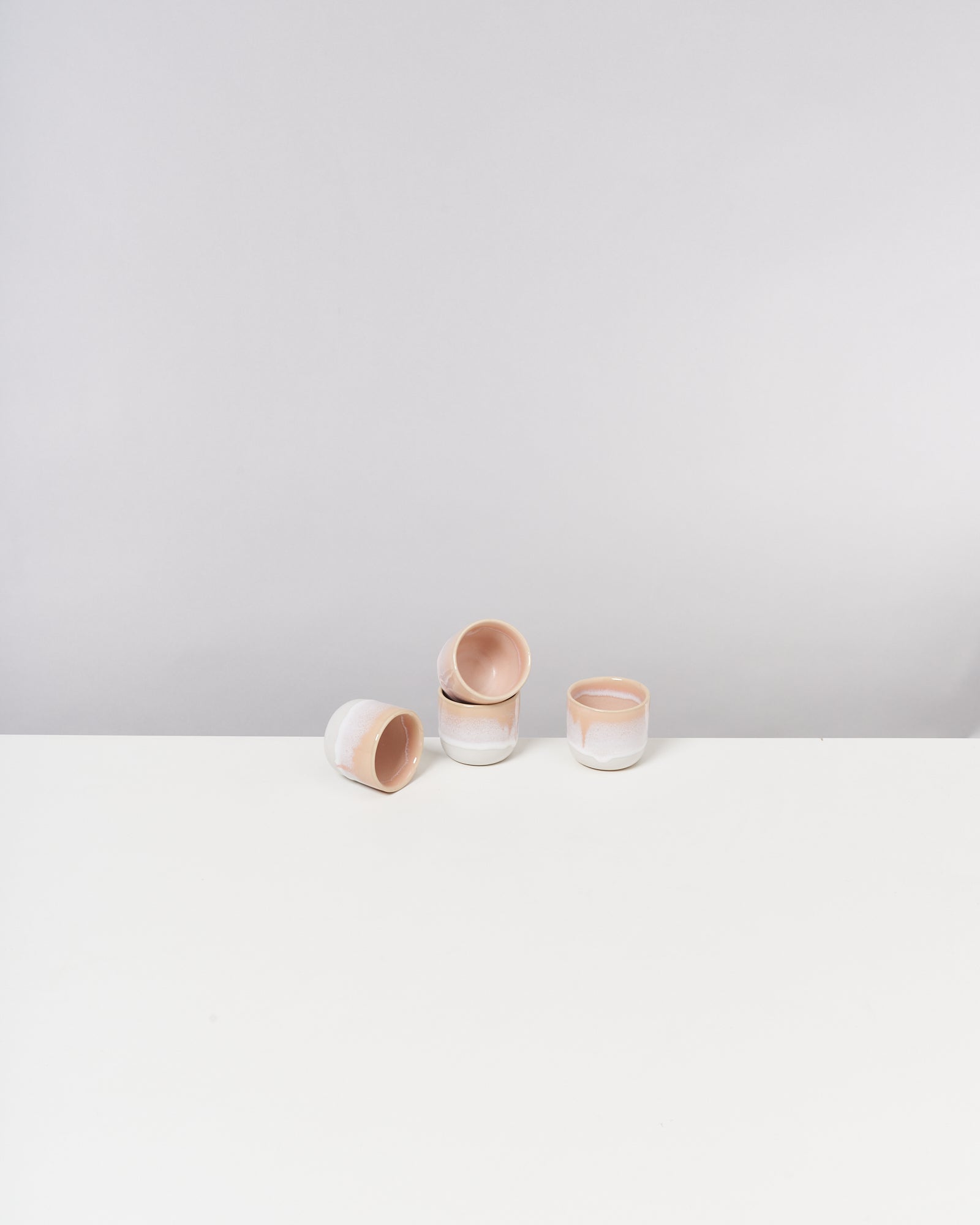 Vier beige-weiße Keramiktassen stehen auf einem weißen Tisch vor einem grauen Hintergrund.