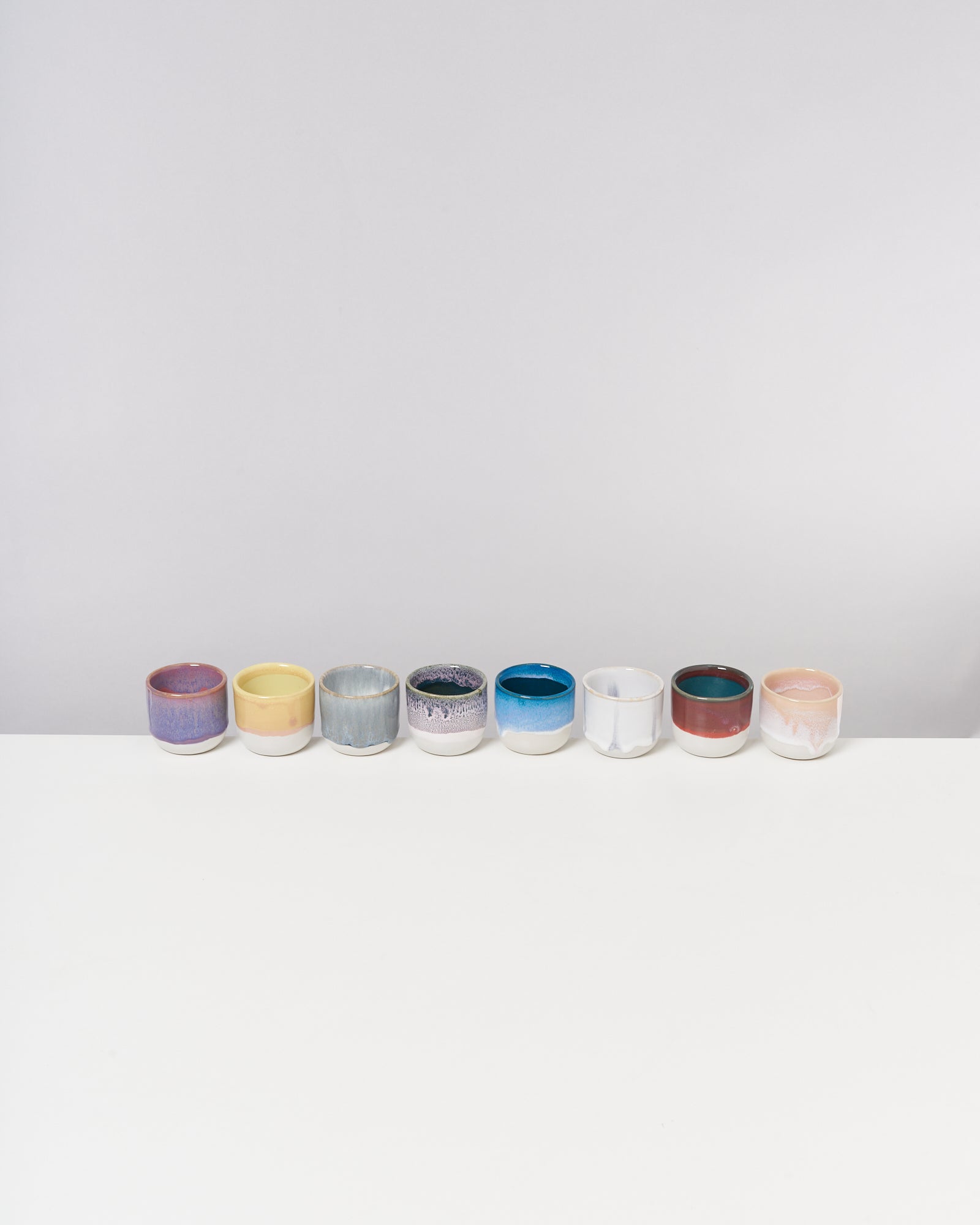 Acht bunte Keramikbecher stehen in einer Reihe auf einem weißen Tisch vor grauem Hintergrund.