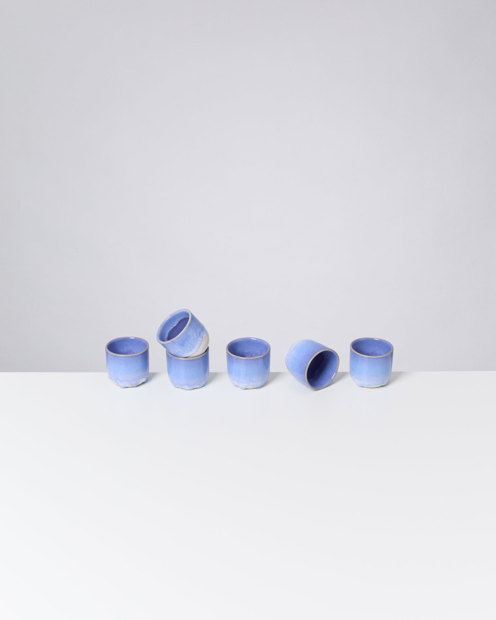 Sechs blaue Keramiktassen stehen und liegen auf einem weißen Tisch vor einem grauen Hintergrund.