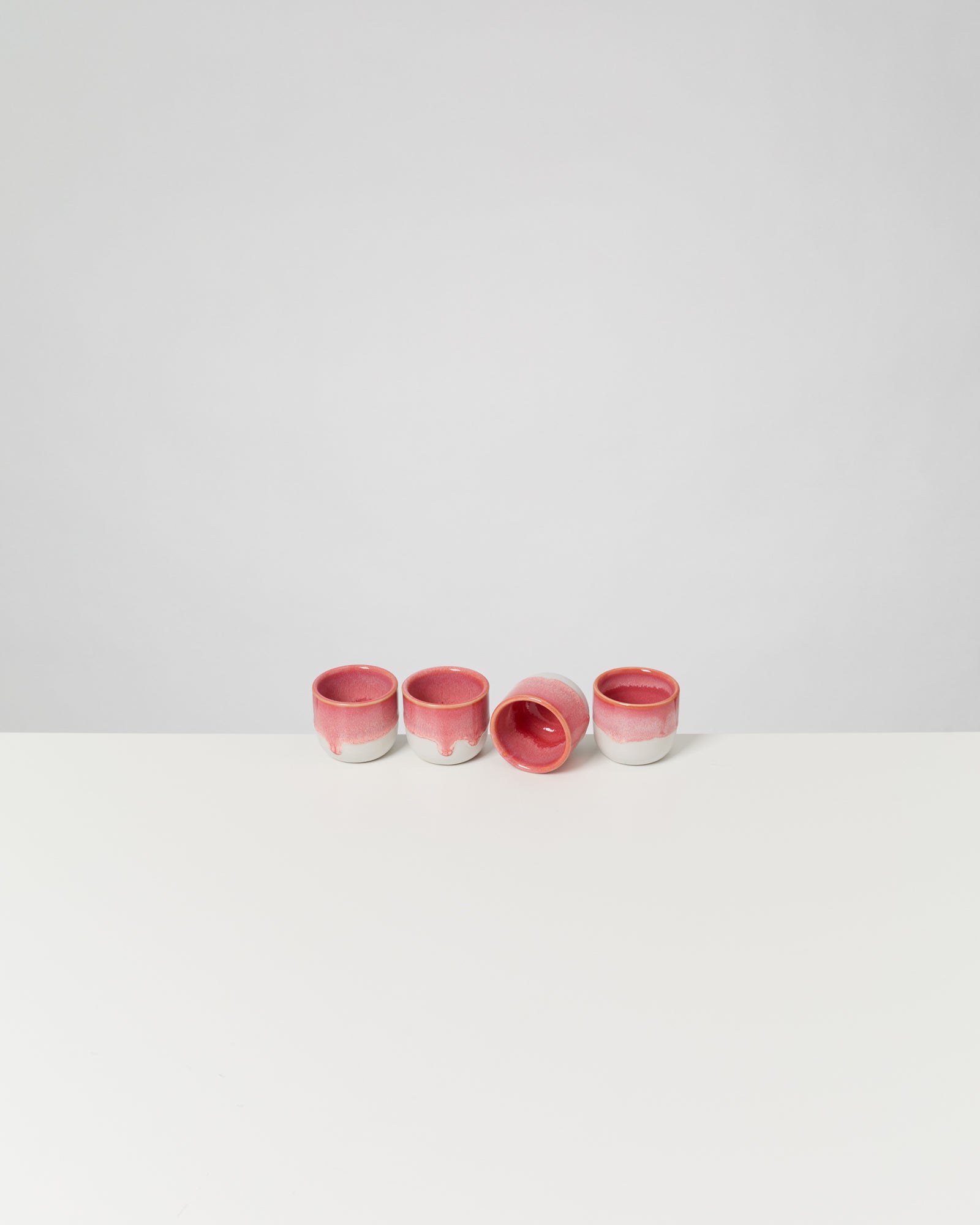 Vier rosa und weiße Keramikbecher, einer liegt auf der Seite, auf einer weißen Fläche vor grauem Hintergrund.