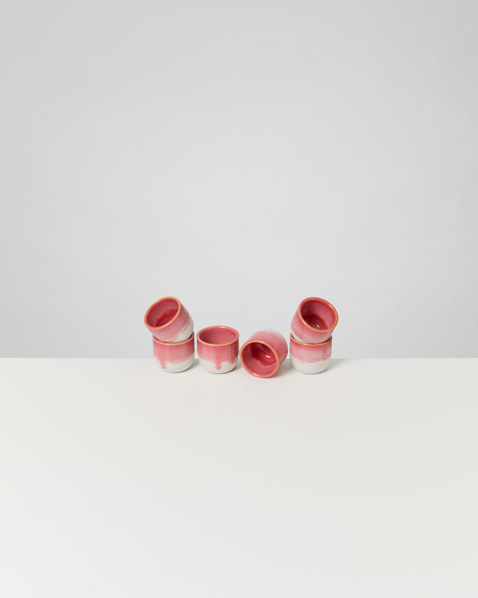 Sechs rosa Becher mit weißem Boden stehen schräg auf einer weißen Oberfläche vor einem grauen Hintergrund.