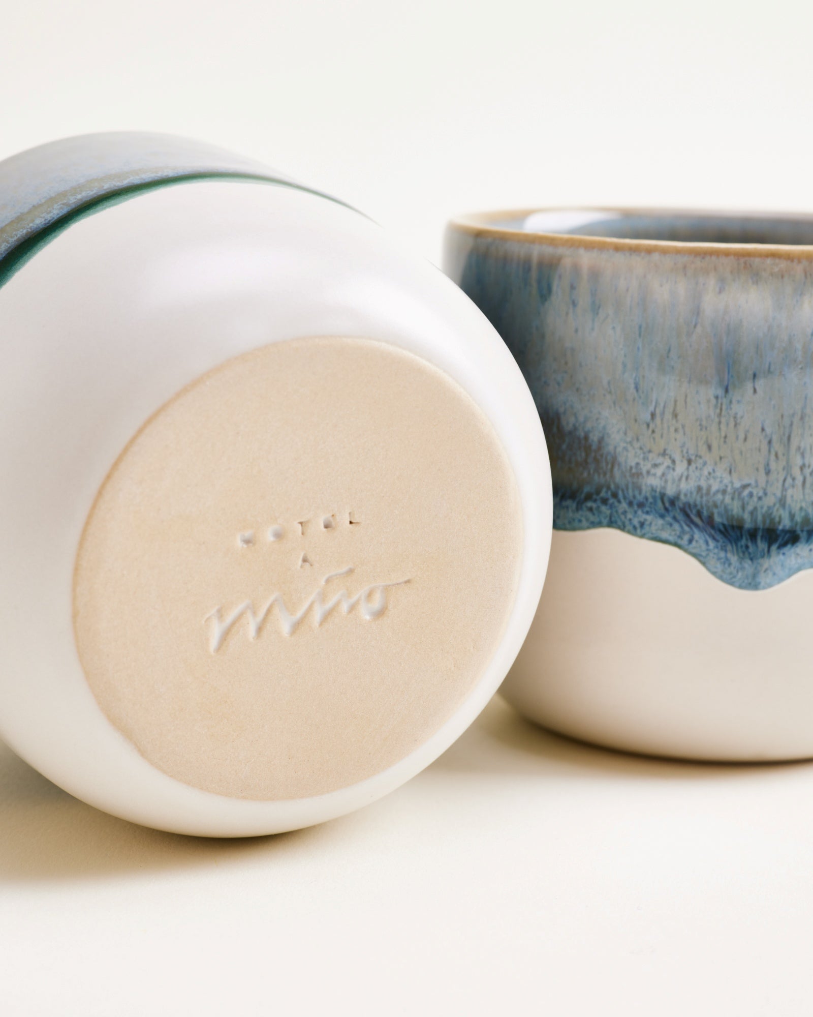 Zwei Keramiktassen mit blauer Glasur, eine liegend, zeigt den Boden mit eingeprägtem Design und Schriftzug.