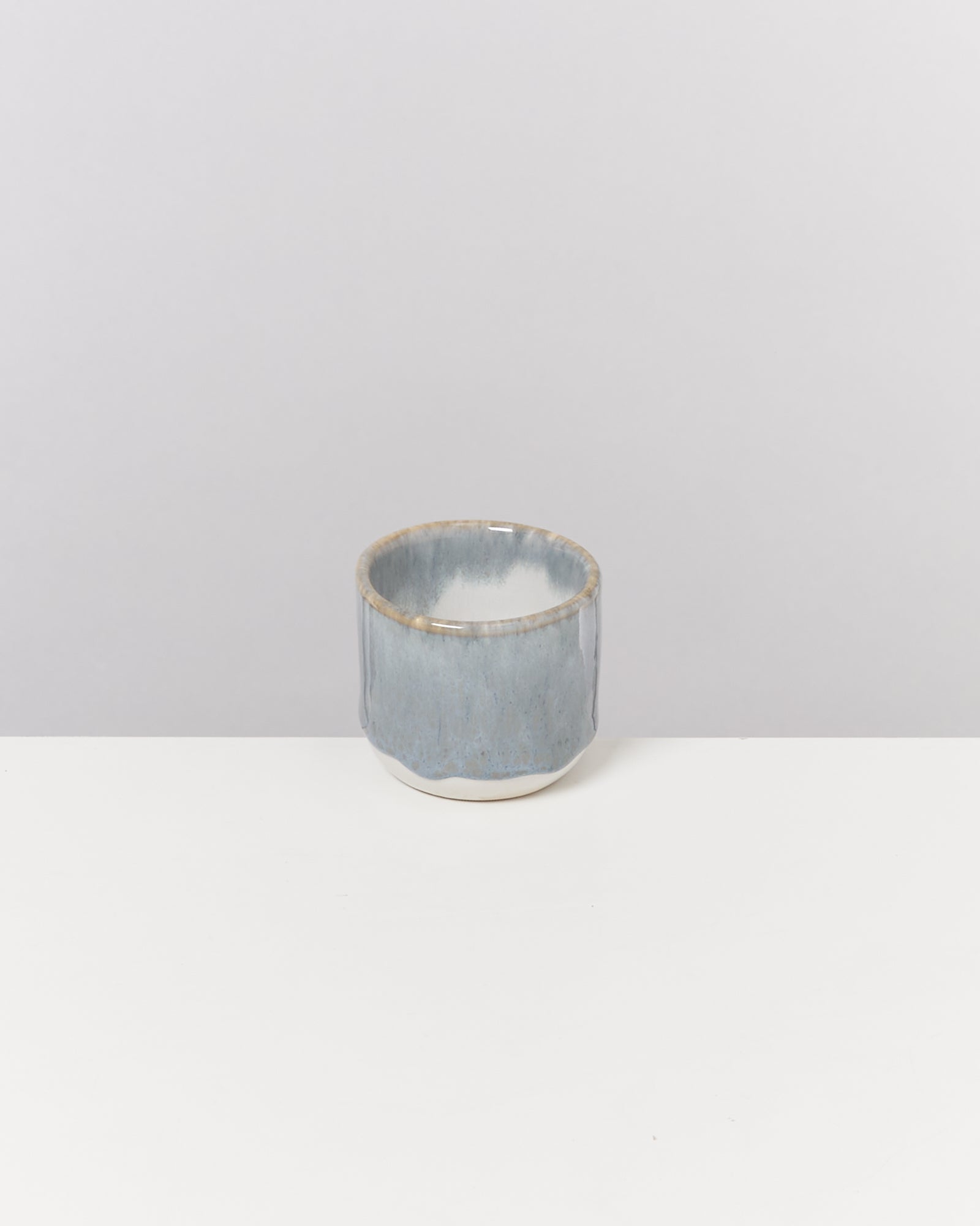 Grauer Keramikbecher mit glattem, glänzendem Finish steht auf einem weißen Tisch vor einem grauen Hintergrund.