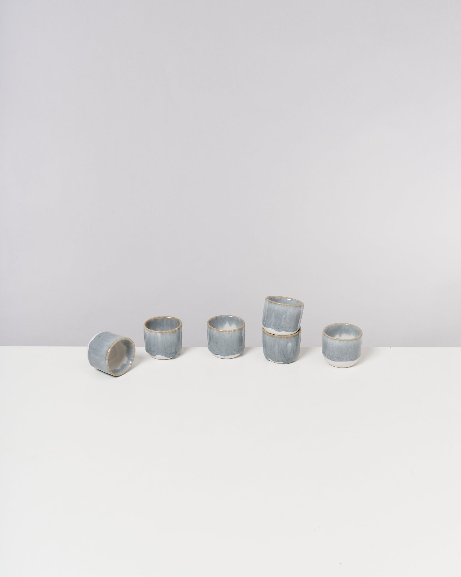 Sechs blaue, handgefertigte Tassen stehen auf einem weißen Tisch, mit einer umgekippten Tasse links.