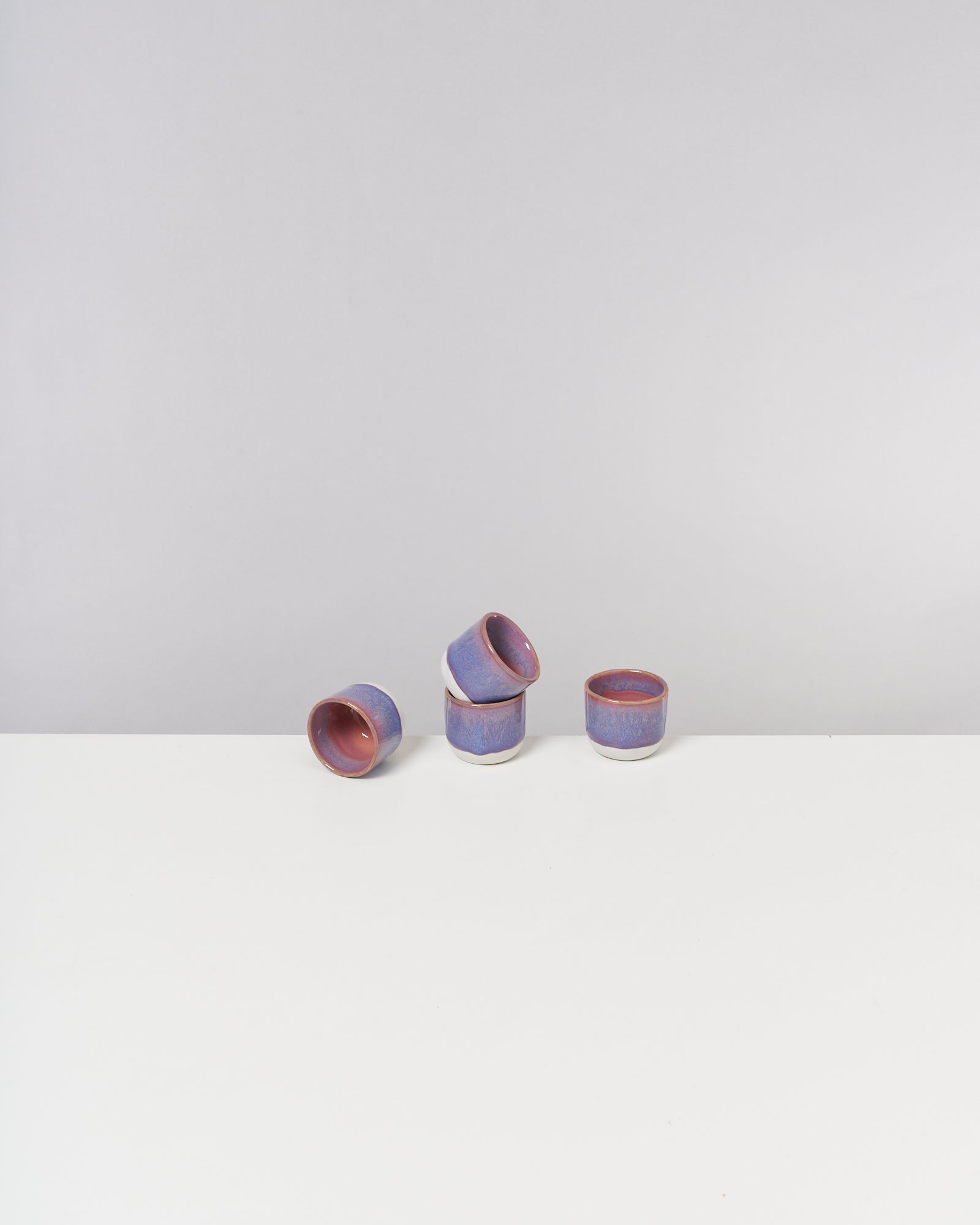 Vier blau-violette Keramiktassen, zwei liegend und zwei aufrecht, auf einem weißen Tisch vor grauem Hintergrund.
