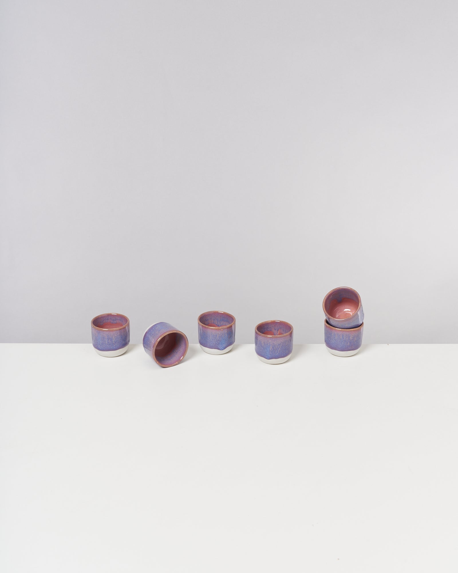 Fünf blau-violette Keramiktassen auf einem weißen Tisch; eine Tasse liegt seitlich.