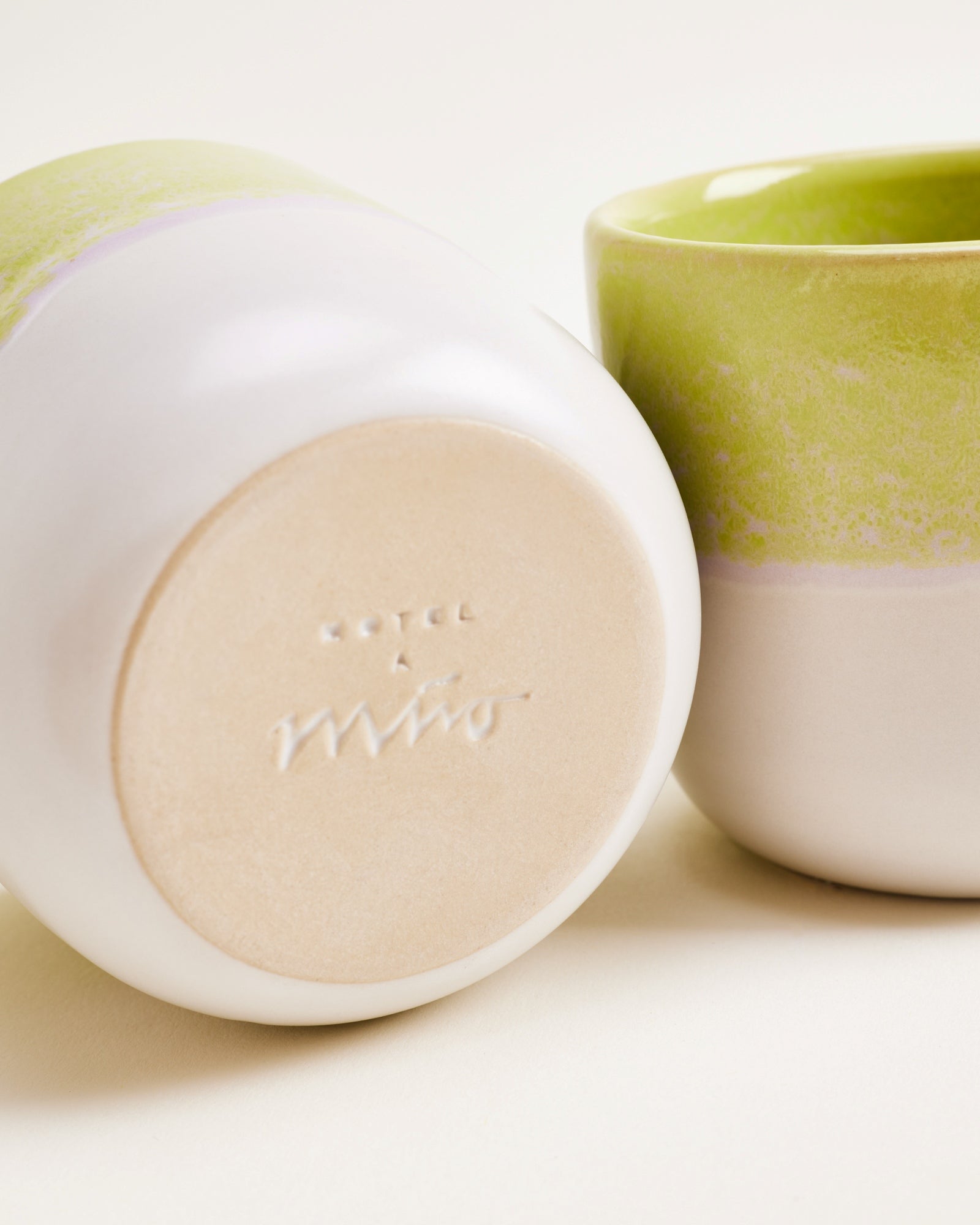 Zwei Keramiktassen, eine auf der Seite liegend, die andere stehend, mit hellgrünem oberen Rand auf hellem Hintergrund.