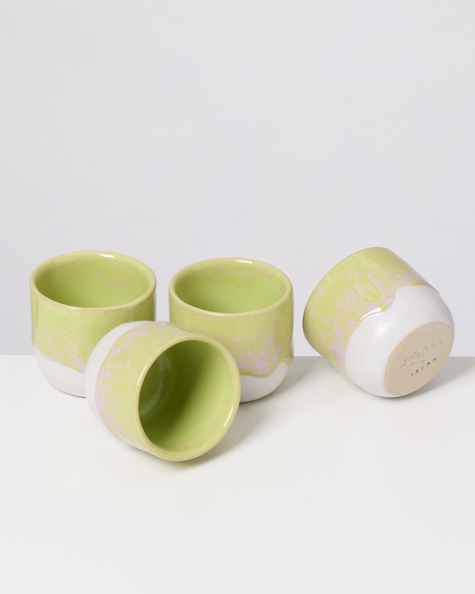 Vier hellgrüne und weiße Keramikbecher liegen auf einer weißen Oberfläche.