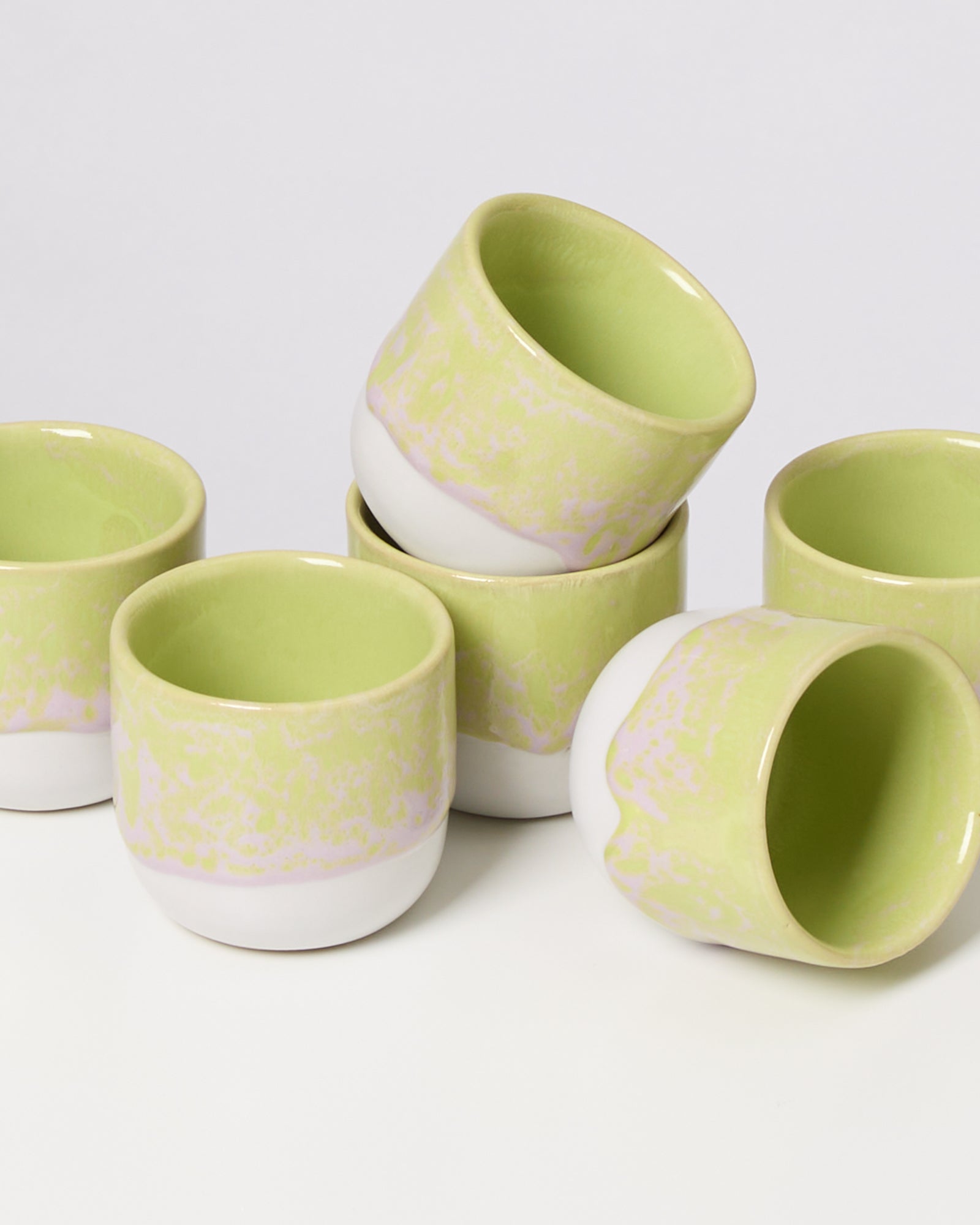 Sechs grüne und weiße Keramiktassen stehen teils gestapelt auf einer weißen Oberfläche.