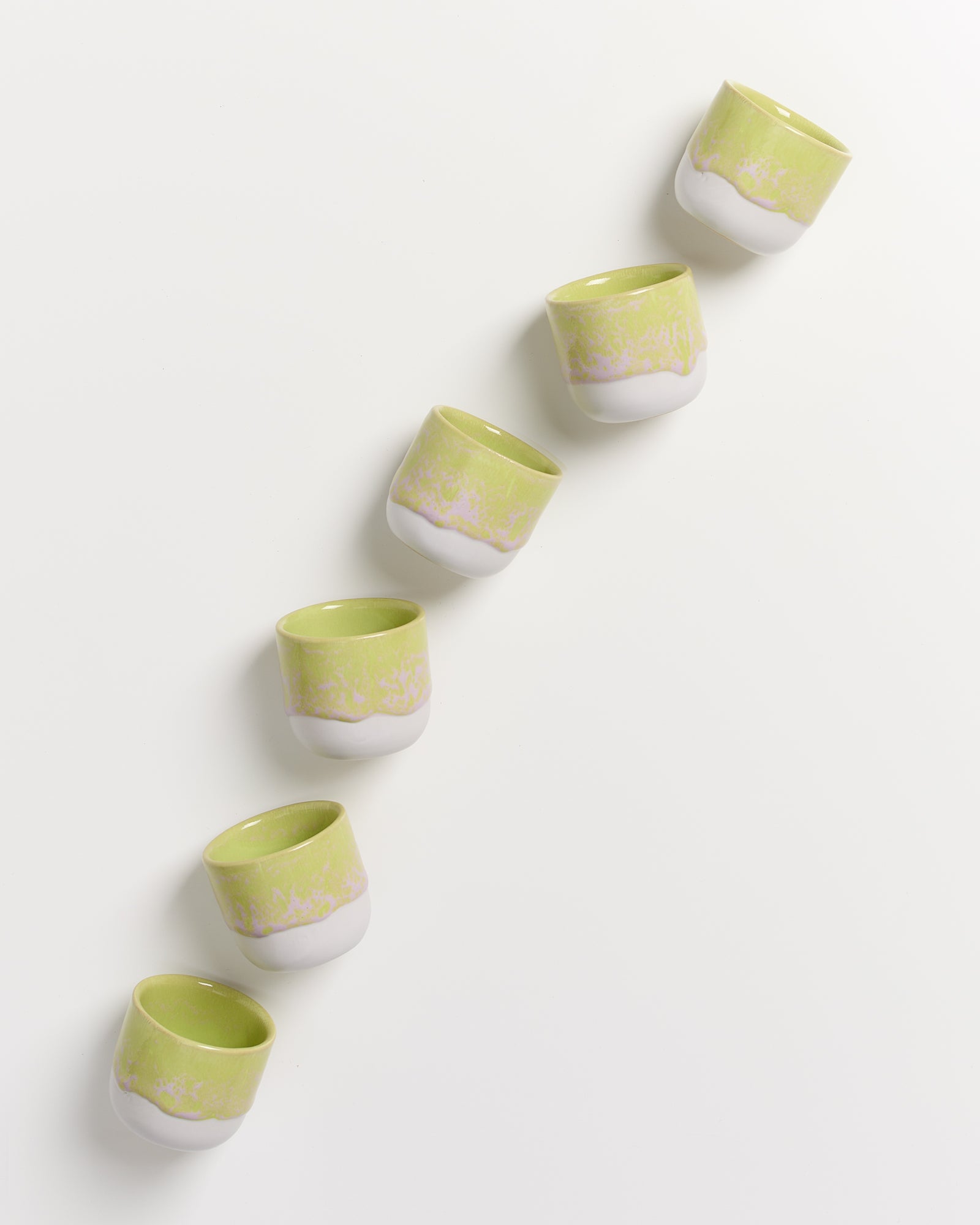 Sechs grün-weiße Keramiktassen sind diagonal auf weißem Hintergrund angeordnet.