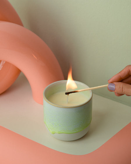 Eine Hand mit lila Nagellack zündet eine Kerze in einem grünen Becher vor einem rosa Hintergrund an.