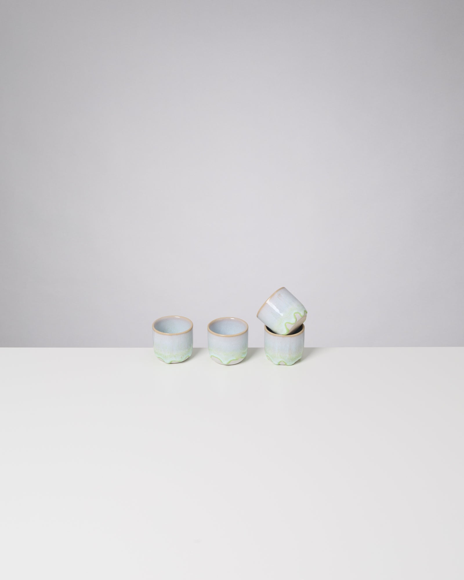 Vier kleine, pastellfarbene Tassen auf einem weißen Tisch vor grauem Hintergrund, eine Tasse liegt schräg auf der anderen.