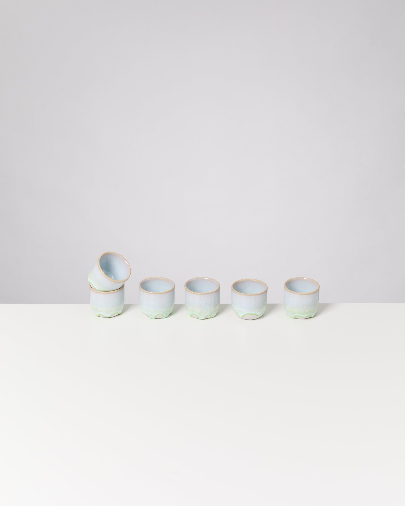 Sechs pastellgrüne Keramiktassen, eine davon auf einer anderen gestapelt, auf einem weißen Tisch vor grauem Hintergrund.