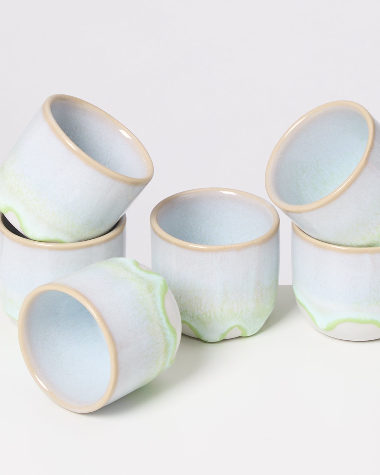 Sechs stapelbare, pastellfarbene Keramiktassen mit leichtem Grünverlauf, auf hellem Hintergrund angeordnet.