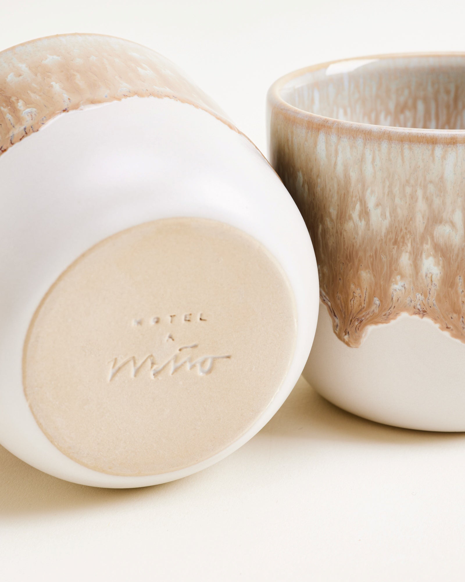Zwei Keramiktassen mit beiger und weißer Glasur, eine liegt auf der Seite und zeigt den Boden mit eingeprägtem Schriftzug.