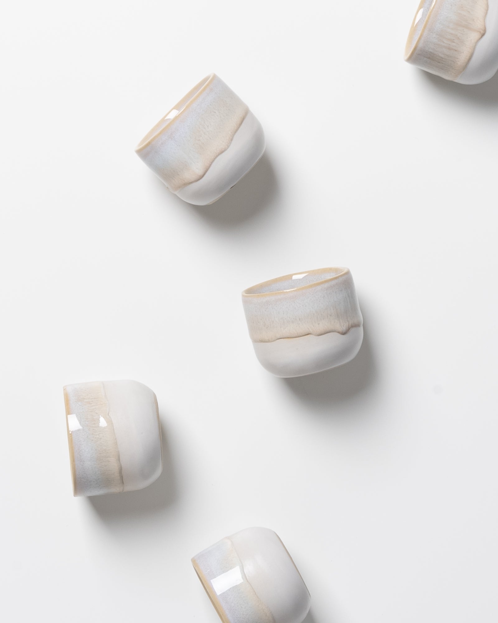 Fünf weiß-beige Keramikbecher auf weißem Hintergrund, ungleichmäßig verteilt.