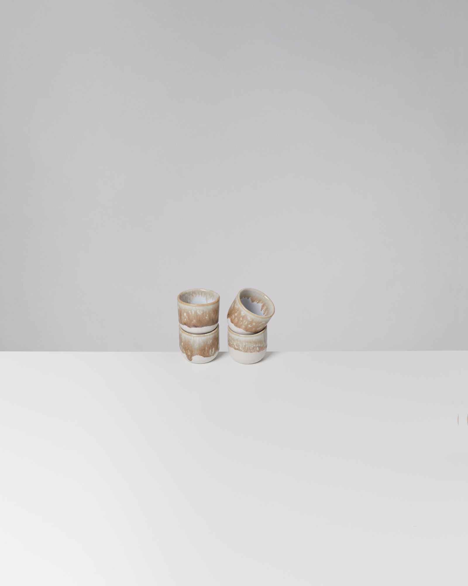 Vier stapelbare, beige-weiße Keramikbecher auf weißem Tisch, minimalistisch vor grauem Hintergrund.