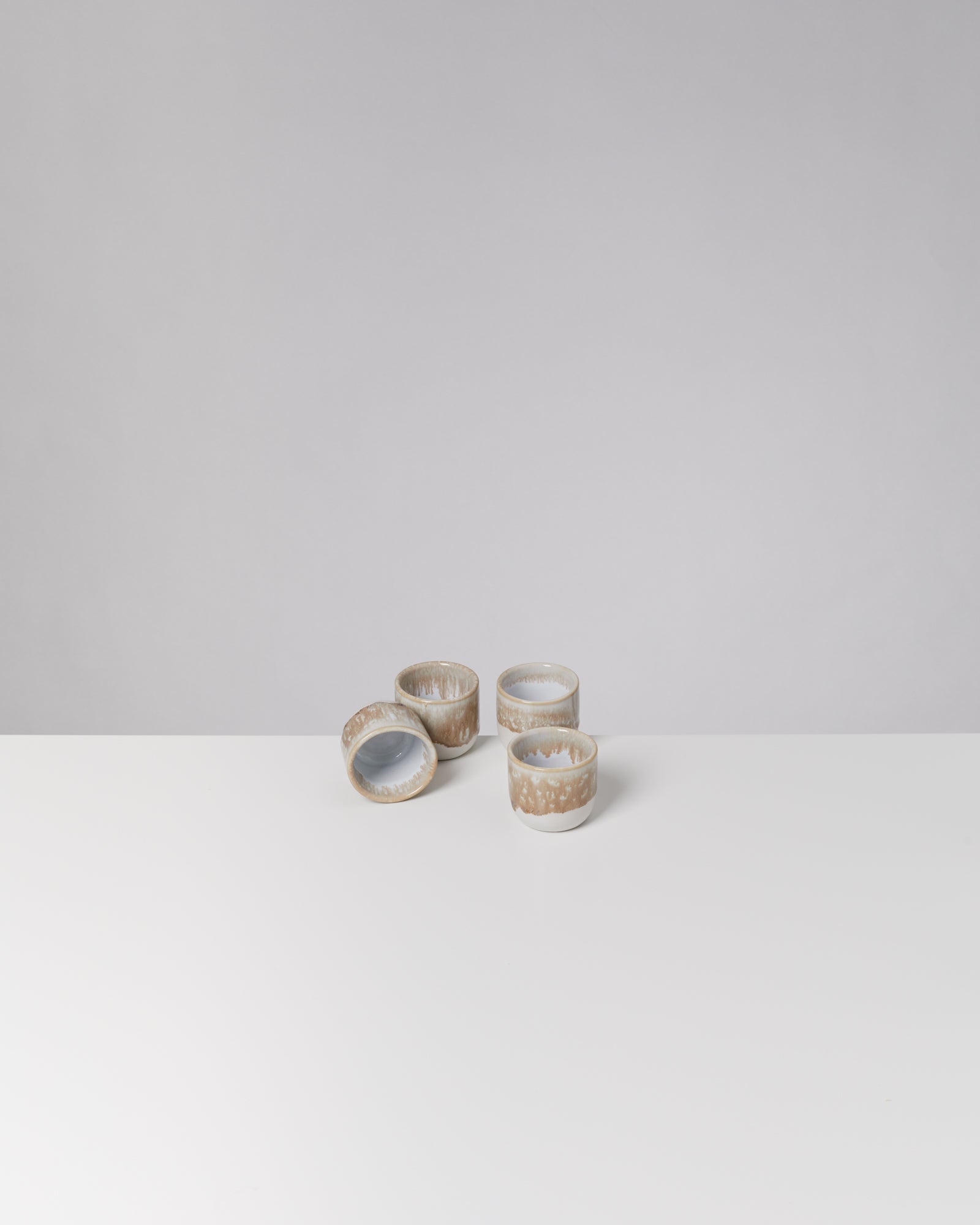 Vier kleine, braun-weiß glasierte Tassen auf einer weißen Oberfläche vor grauem Hintergrund.