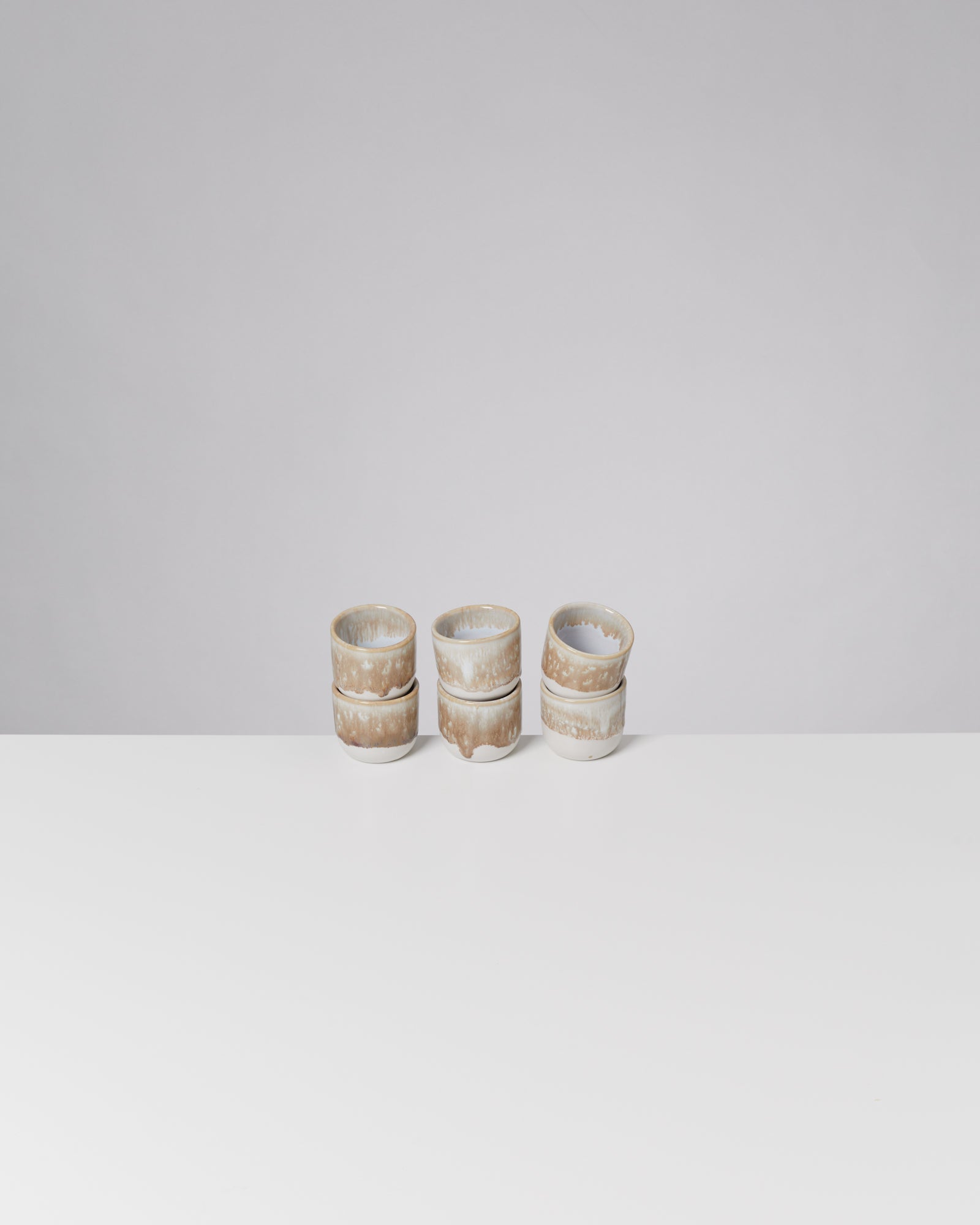 Drei gestapelte, beige-weiße Keramikbecher stehen auf einem weißen Tisch vor grauem Hintergrund.