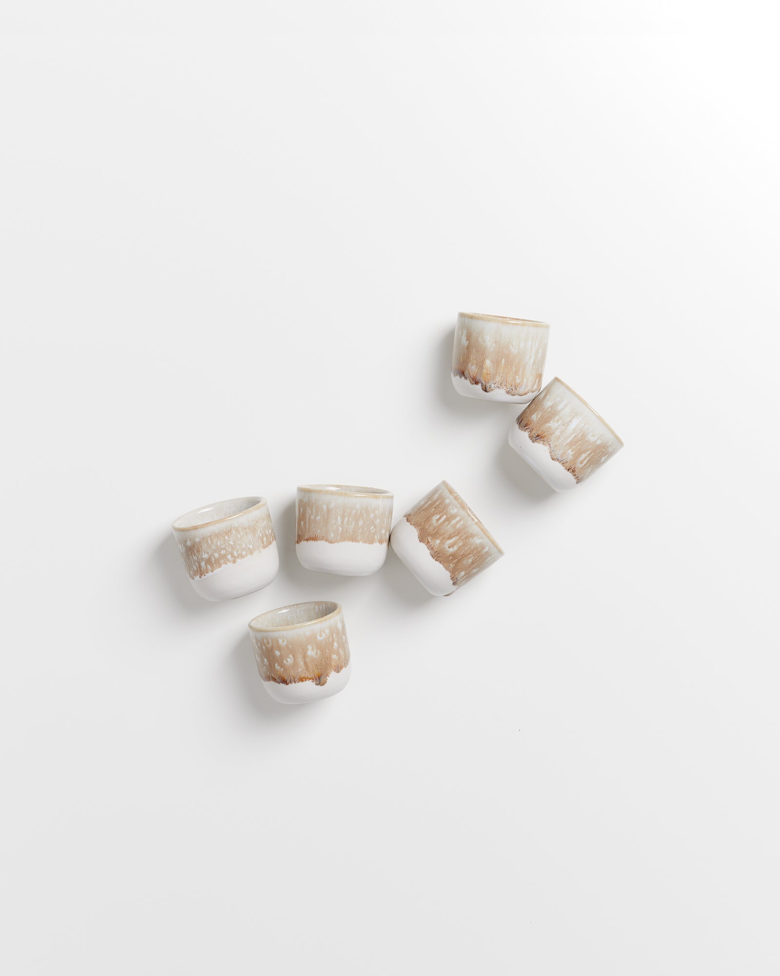 Sechs braun-weiß gemusterte Keramikbecher liegen ungleichmäßig auf einer weißen Oberfläche verteilt.