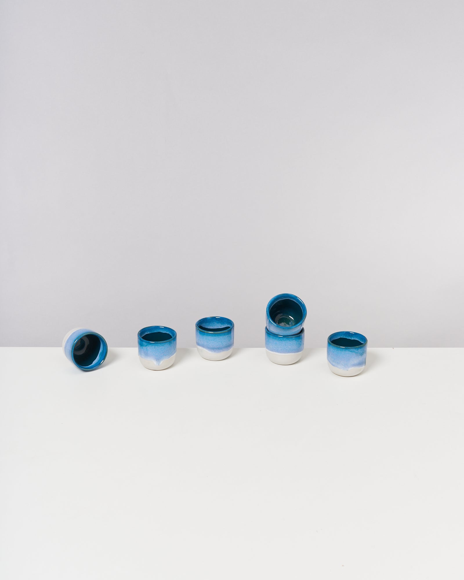 Sechs blau-weiße Keramiktassen stehen und liegen auf einem weißen Tisch vor einem grauen Hintergrund.