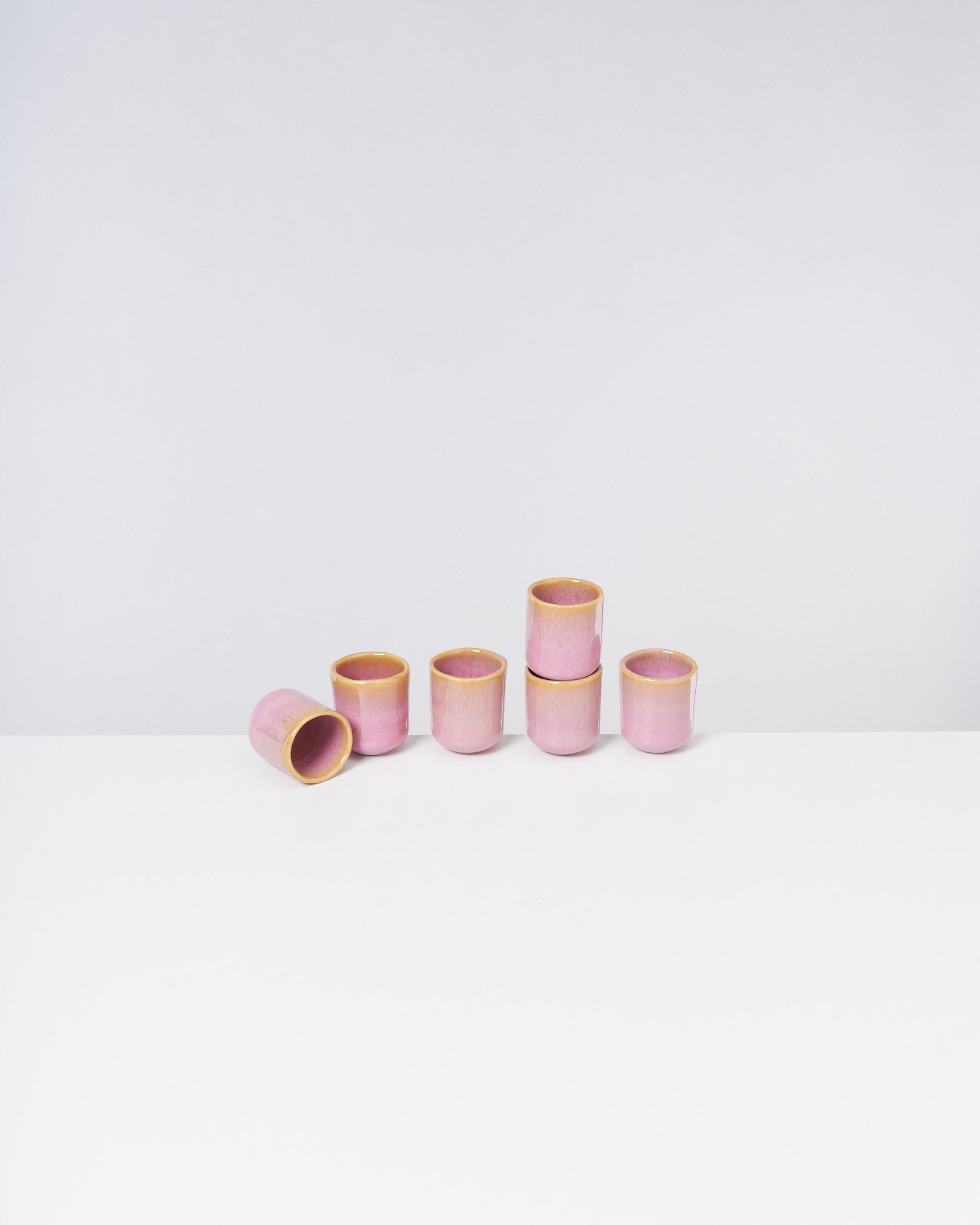 Sechs rosa Keramiktassen, eine liegt auf der Seite, auf einer weißen Oberfläche vor grauem Hintergrund.
