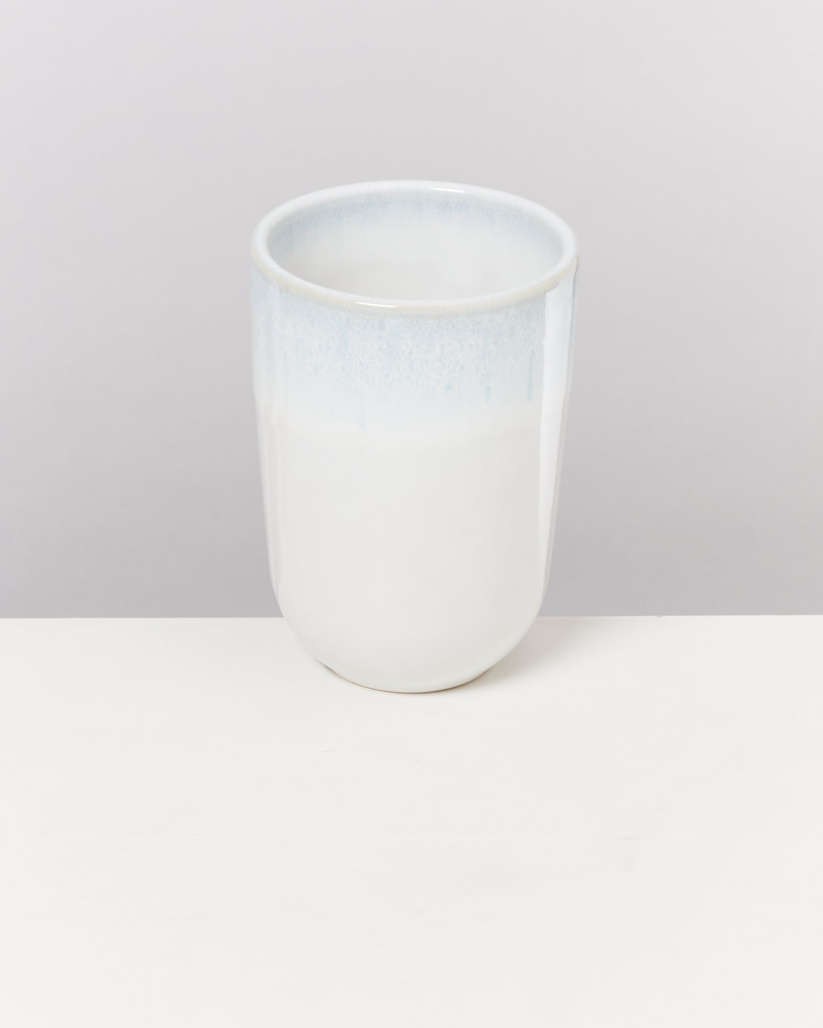 Weißer Keramikbecher mit blauer Oberkante auf einem weißen Tisch vor grauem Hintergrund.