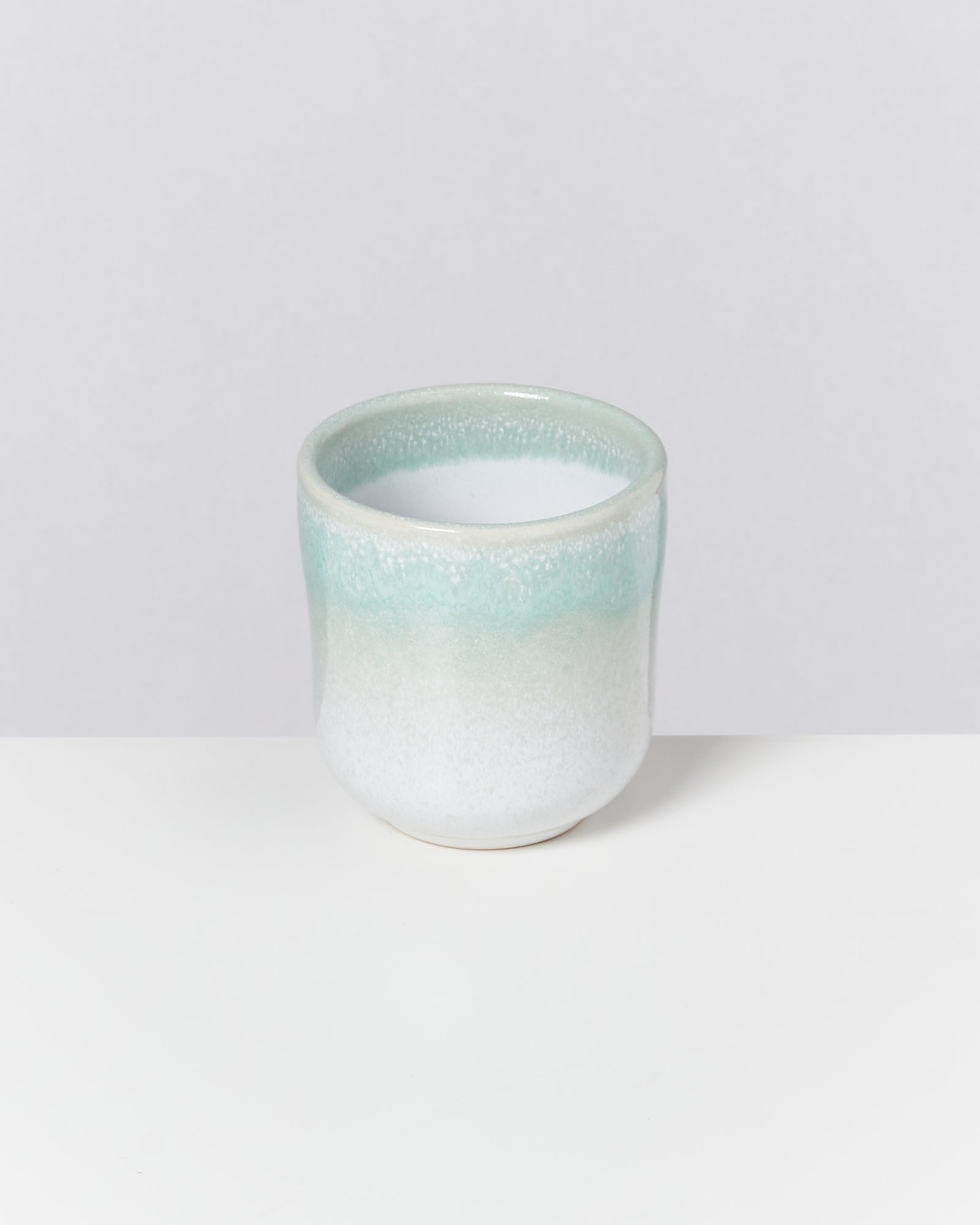 Ein kleiner, zylindrischer Keramikbecher in Pastellgrün und Weiß auf einer weißen Oberfläche.