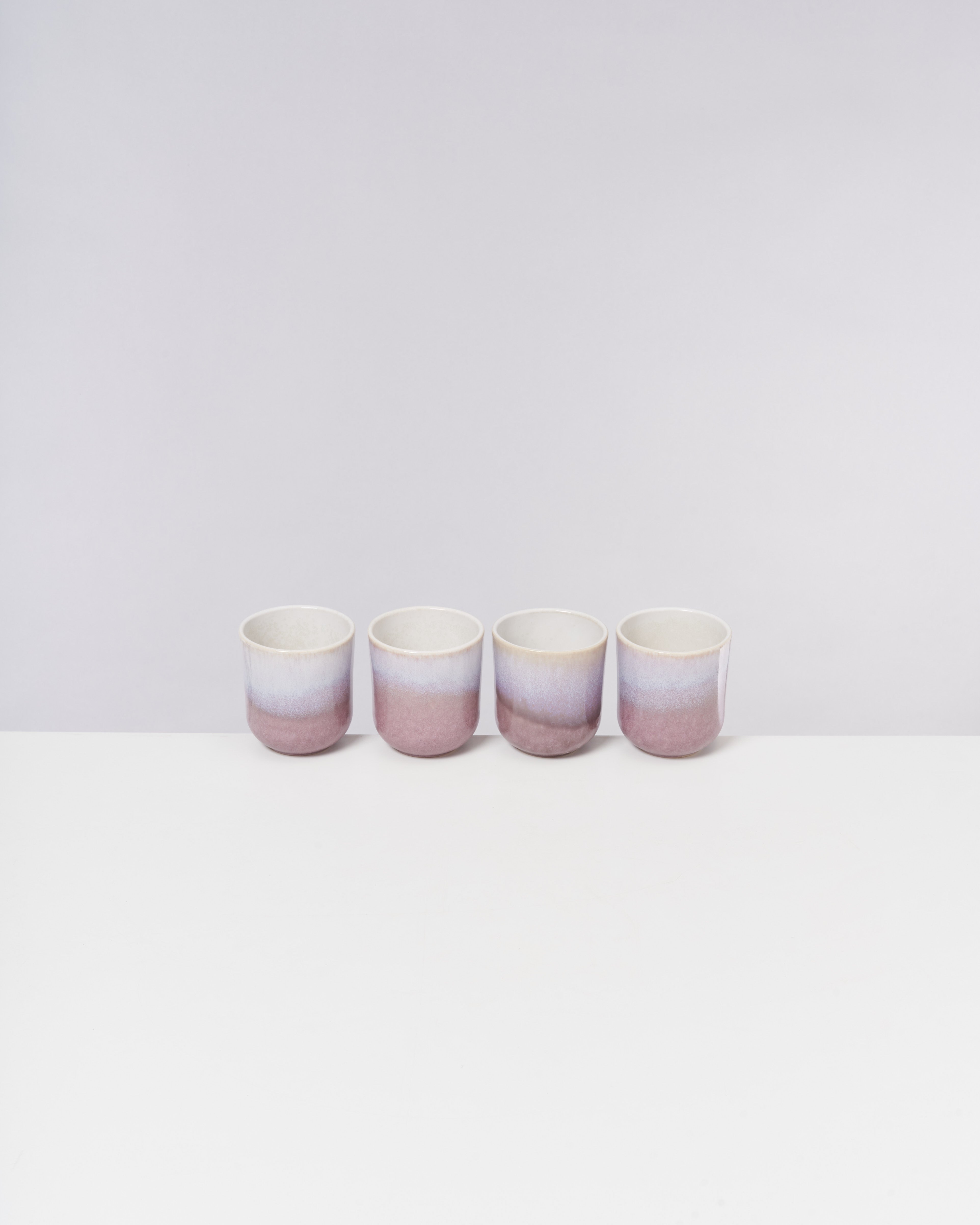 Vier bunte Keramikbecher stehen in einer Reihe auf einem weißen Tisch vor einem hellgrauen Hintergrund.