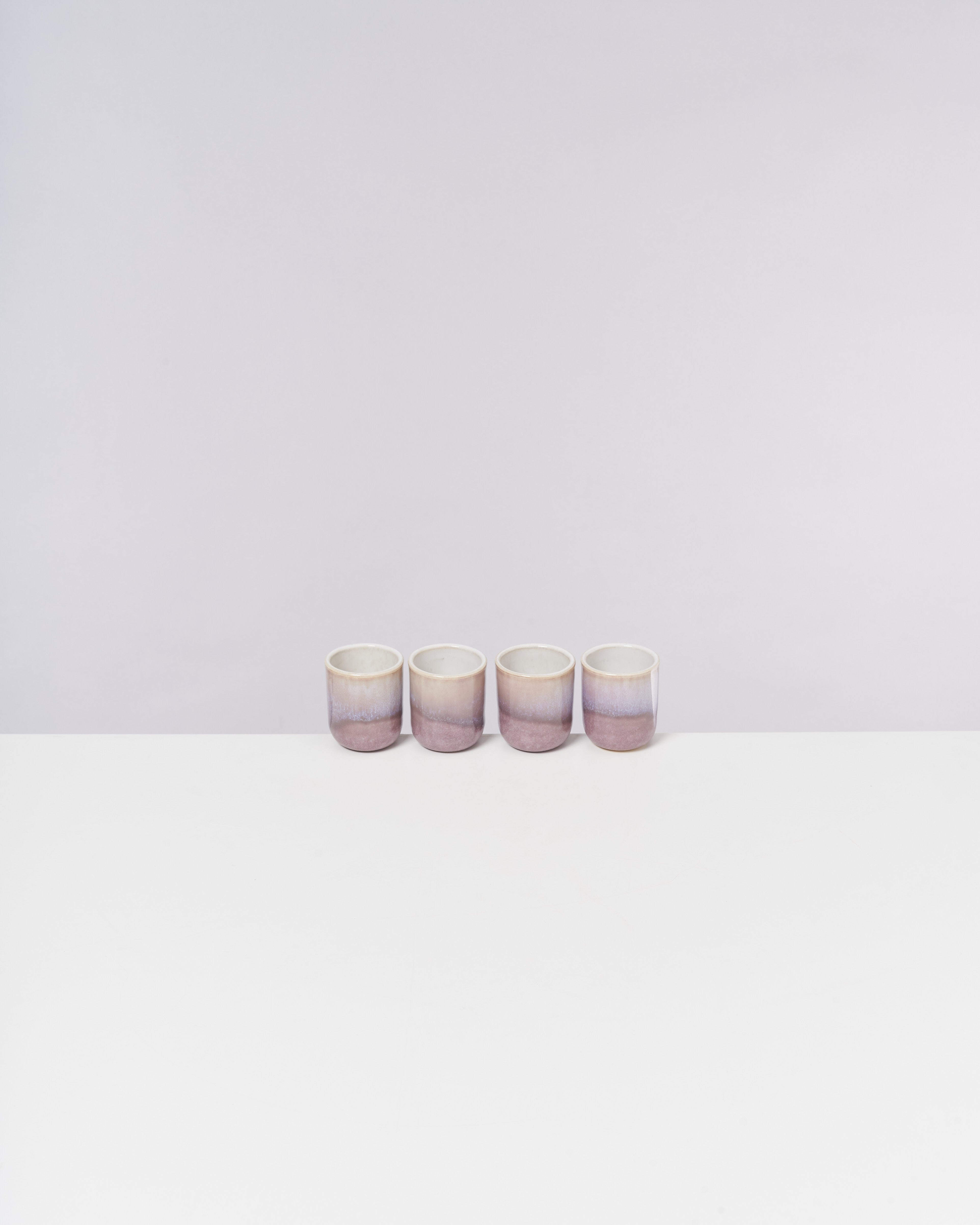 Vier pastellfarbene Tassen stehen nebeneinander auf einer weißen Fläche vor einem hellen Hintergrund.