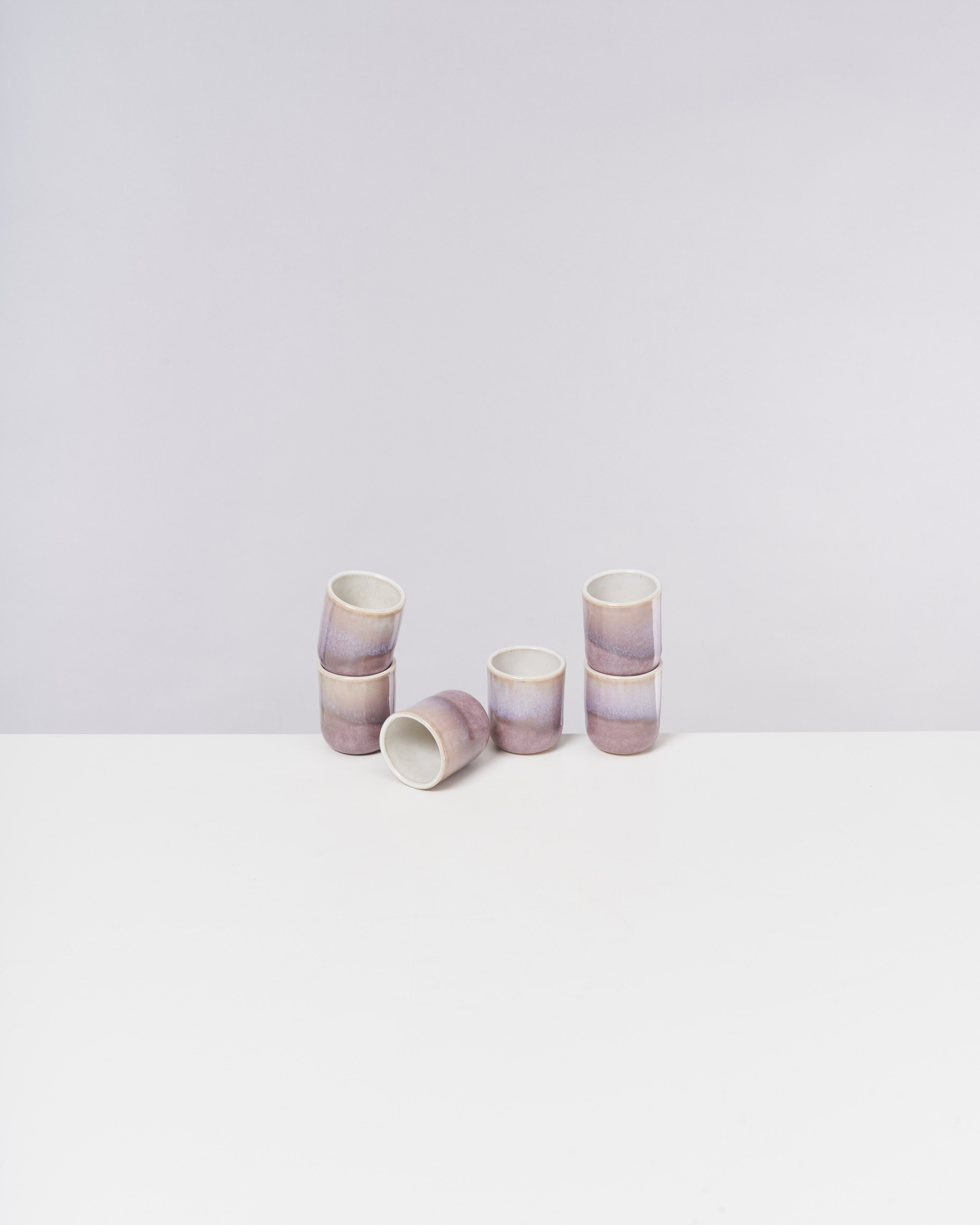 Sechs pastellfarbene Becher, vier aufrecht und zwei liegend, stehen auf einer weißen Oberfläche vor einem hellgrauen Hintergrund.