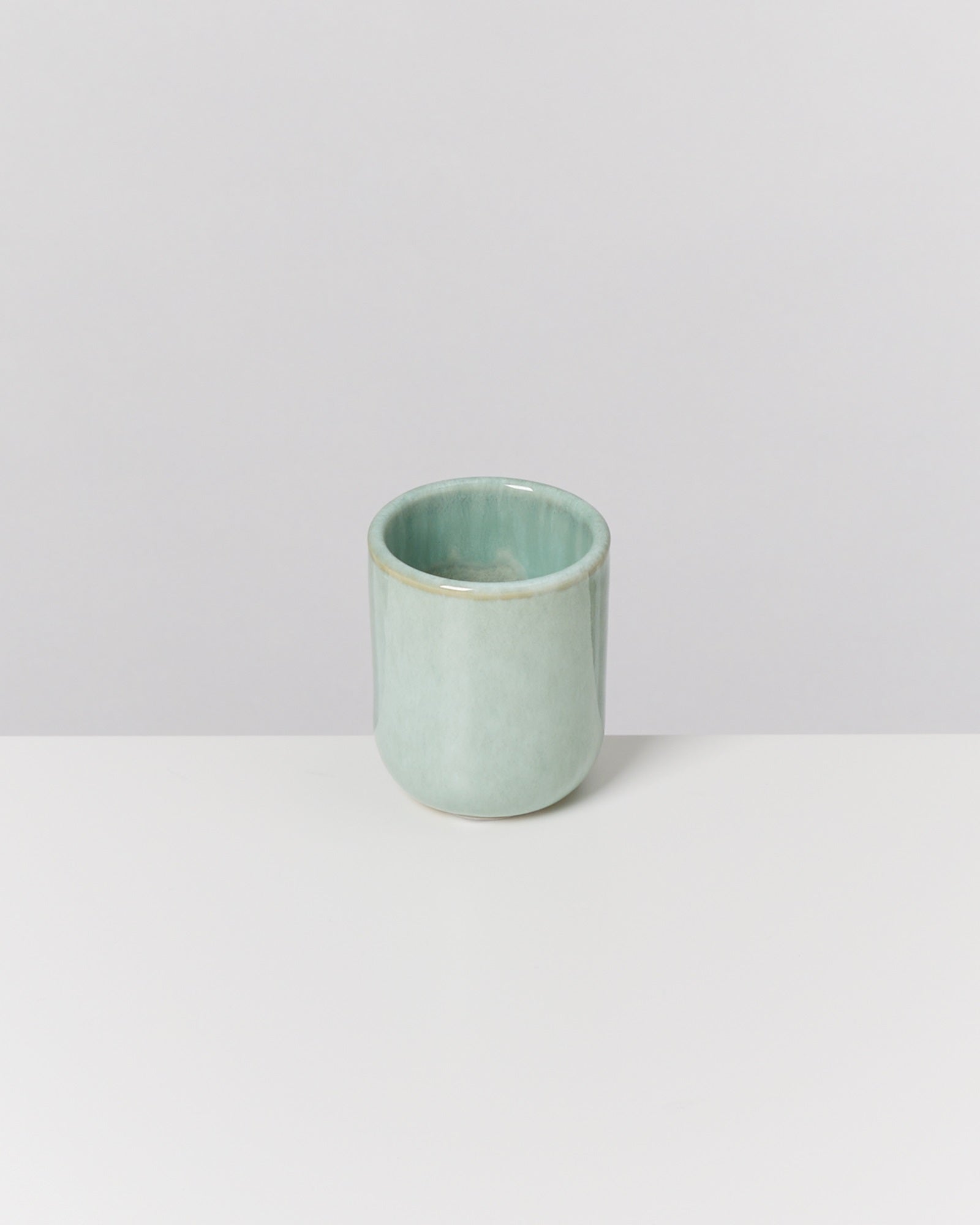 Ein hellgrüner Keramikbecher steht auf einer weißen Oberfläche vor einem grauen Hintergrund.