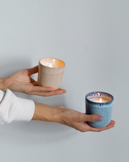 Zwei Hände halten jeweils eine brennende Kerze in pastellfarbenen Keramikgefäßen vor einem neutralen Hintergrund.