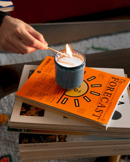 Eine Person zündet eine Kerze auf einem Stapel Bücher mit einem Streichholz an, das oberste Buch hat ein oranges Cover.