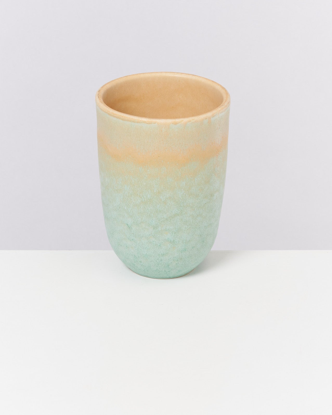 Ein pastellfarbener Keramikbecher mit orange-grünem Farbverlauf steht auf einem weißen Tisch vor grauem Hintergrund.