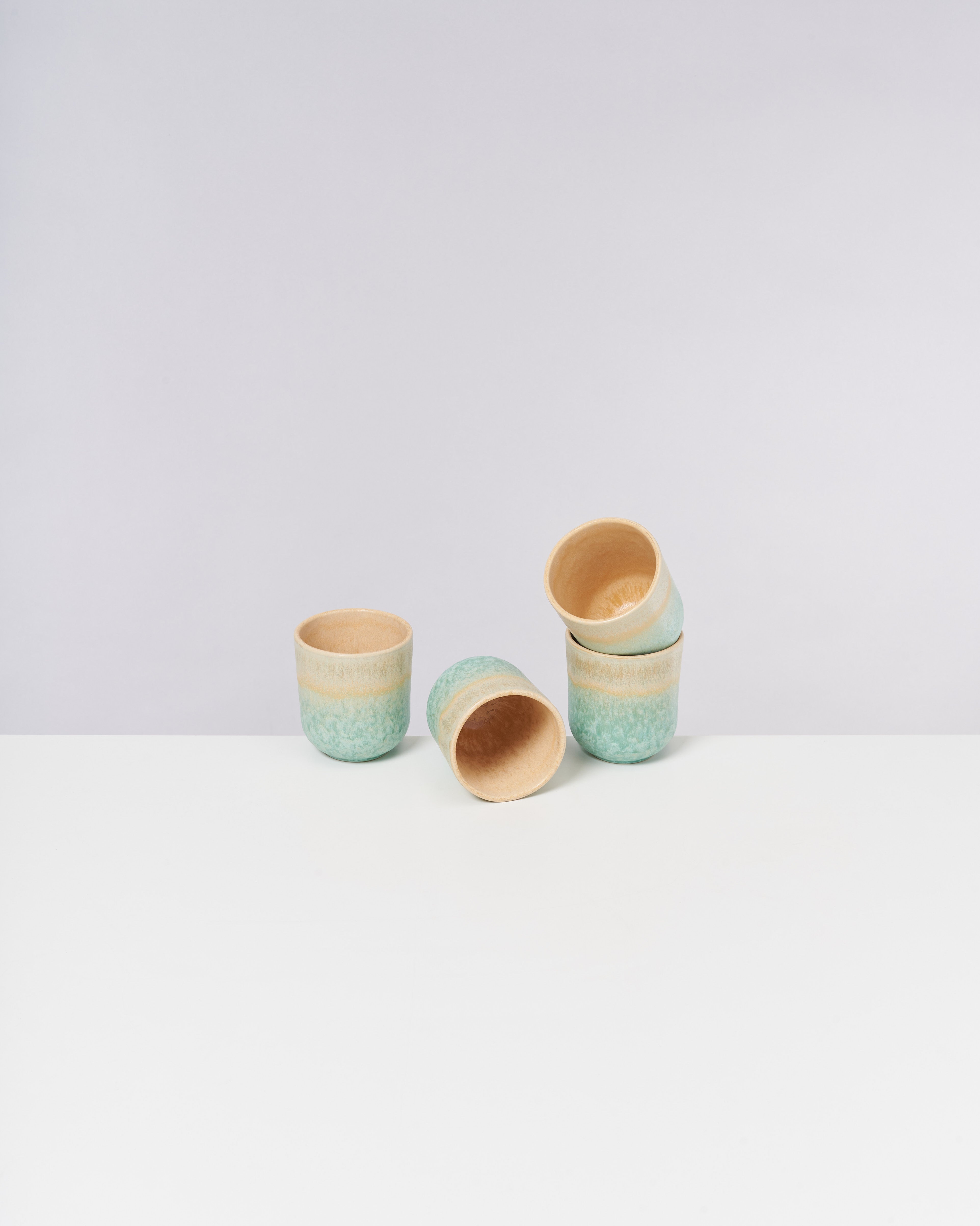 Vier grüne Keramikbecher auf einem weißen Tisch, zwei stehend, einer liegend und einer schräg auf den anderen gestapelt.