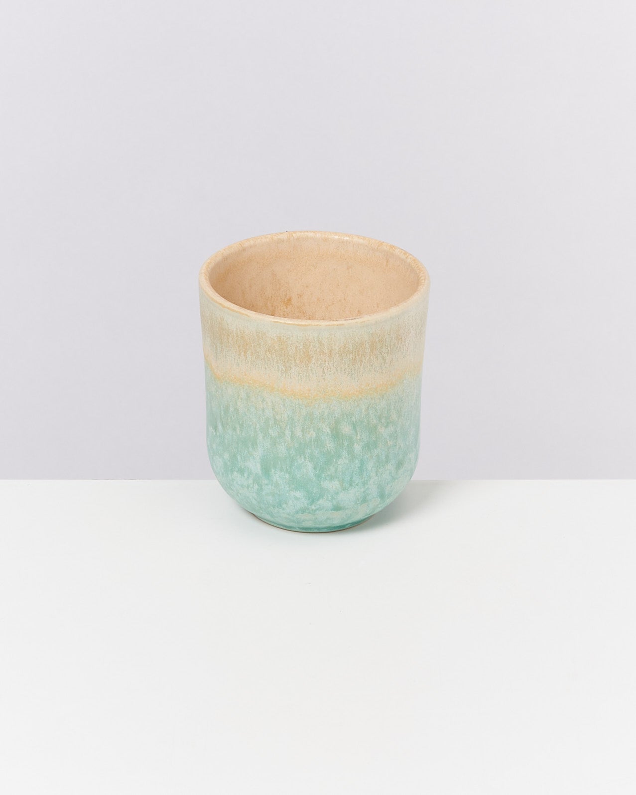 Keramiktasse mit blauer und beiger Glasur auf einem weißen Tisch vor hellem Hintergrund.