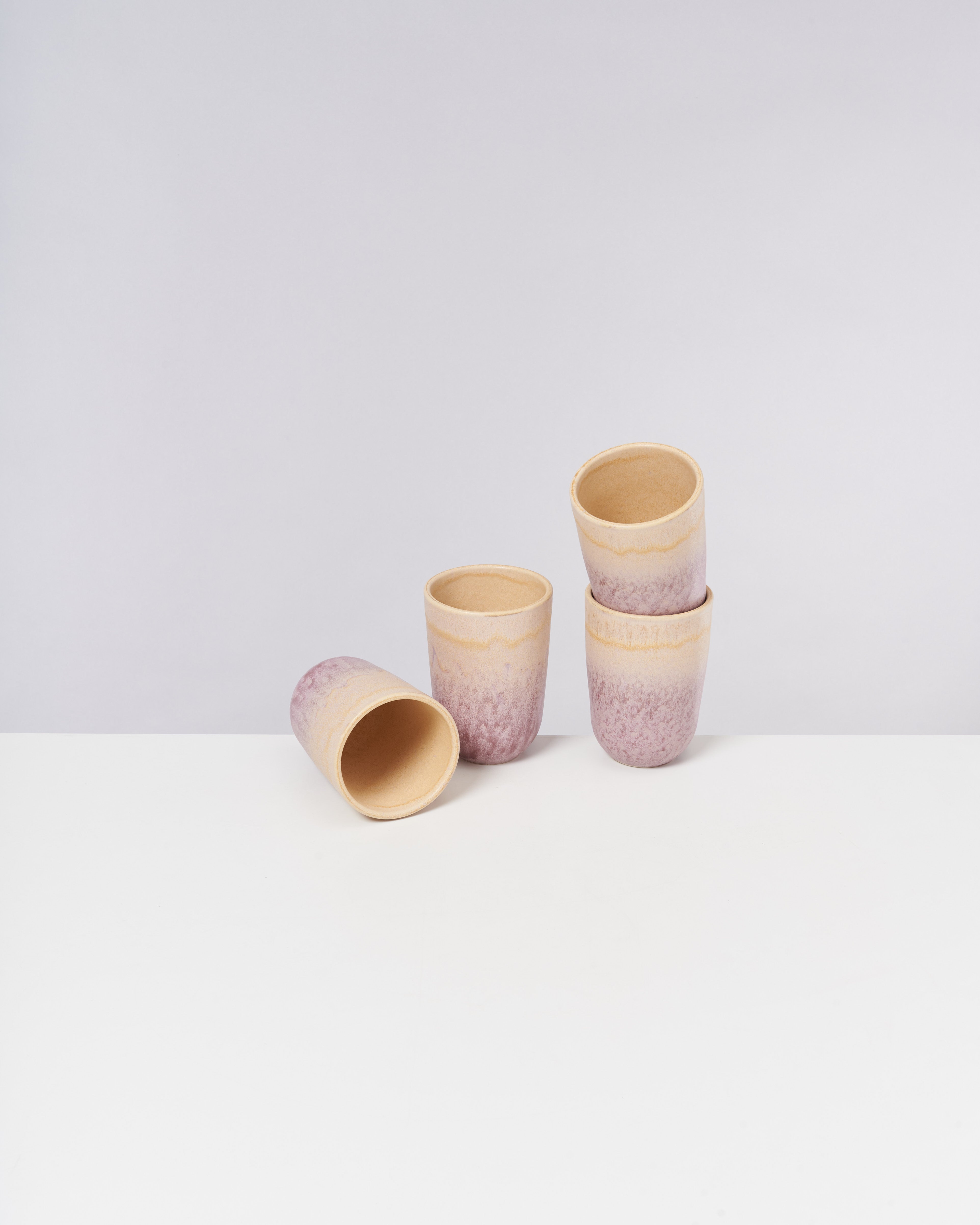 Vier pastellfarbene Keramikbecher, drei stapelbar und einer liegend, auf einem weißen Tisch vor grauem Hintergrund.