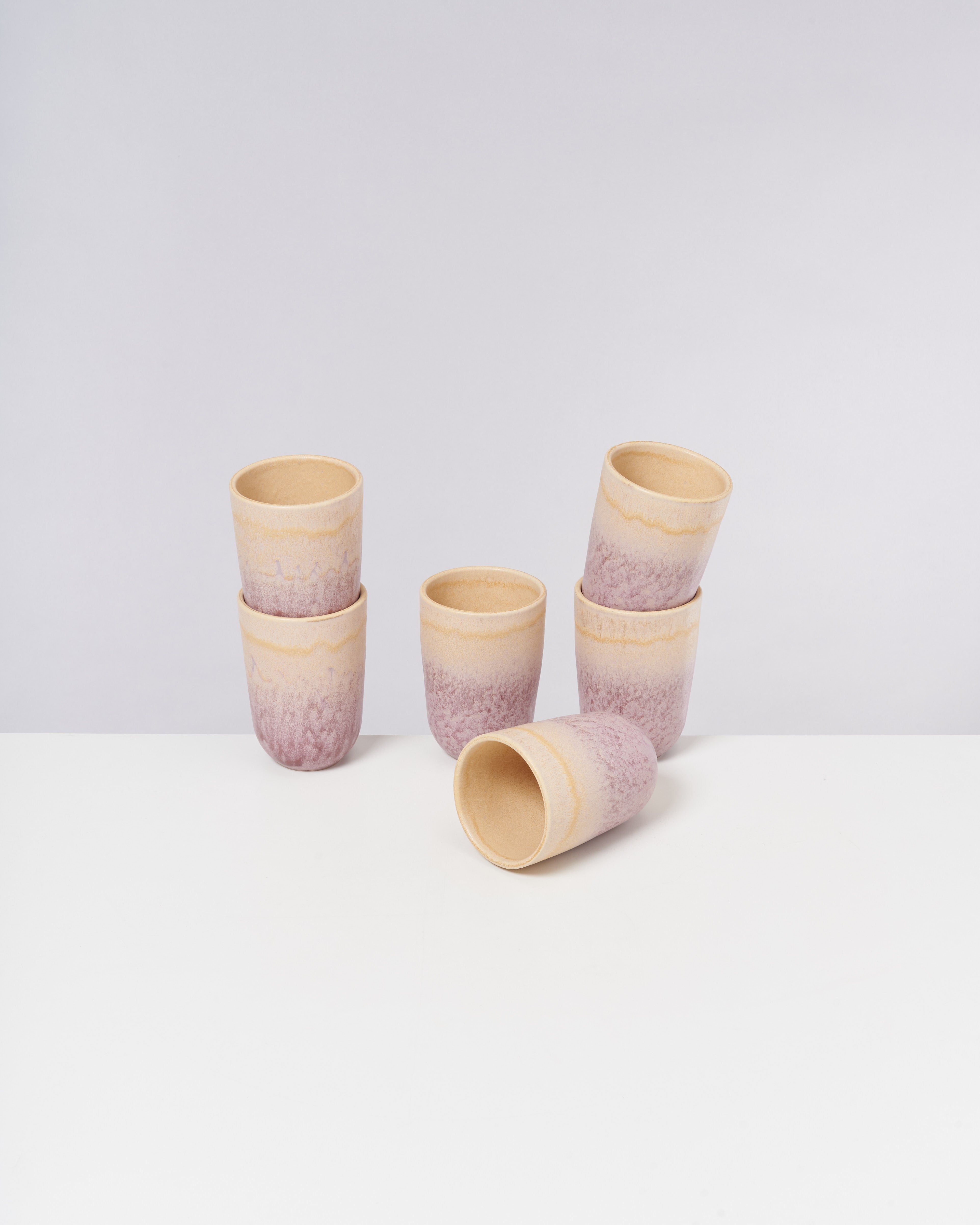 Sechs becherförmige Keramiktassen in hellem Beige und Lila sind auf einem weißen Tisch arrangiert, eine davon liegt seitlich.
