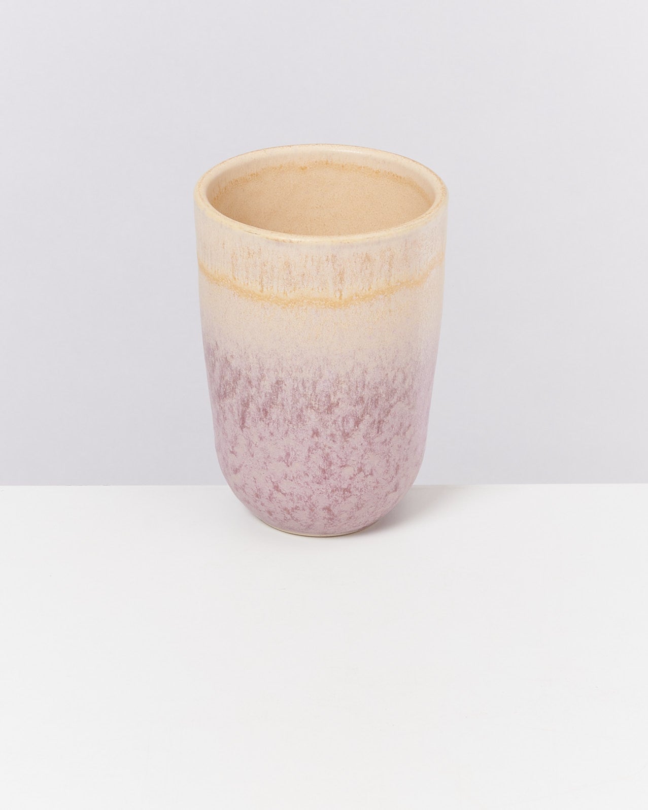 Beiger Keramikbecher mit rosa Farbverlauf steht auf einer weißen Oberfläche vor hellem Hintergrund.