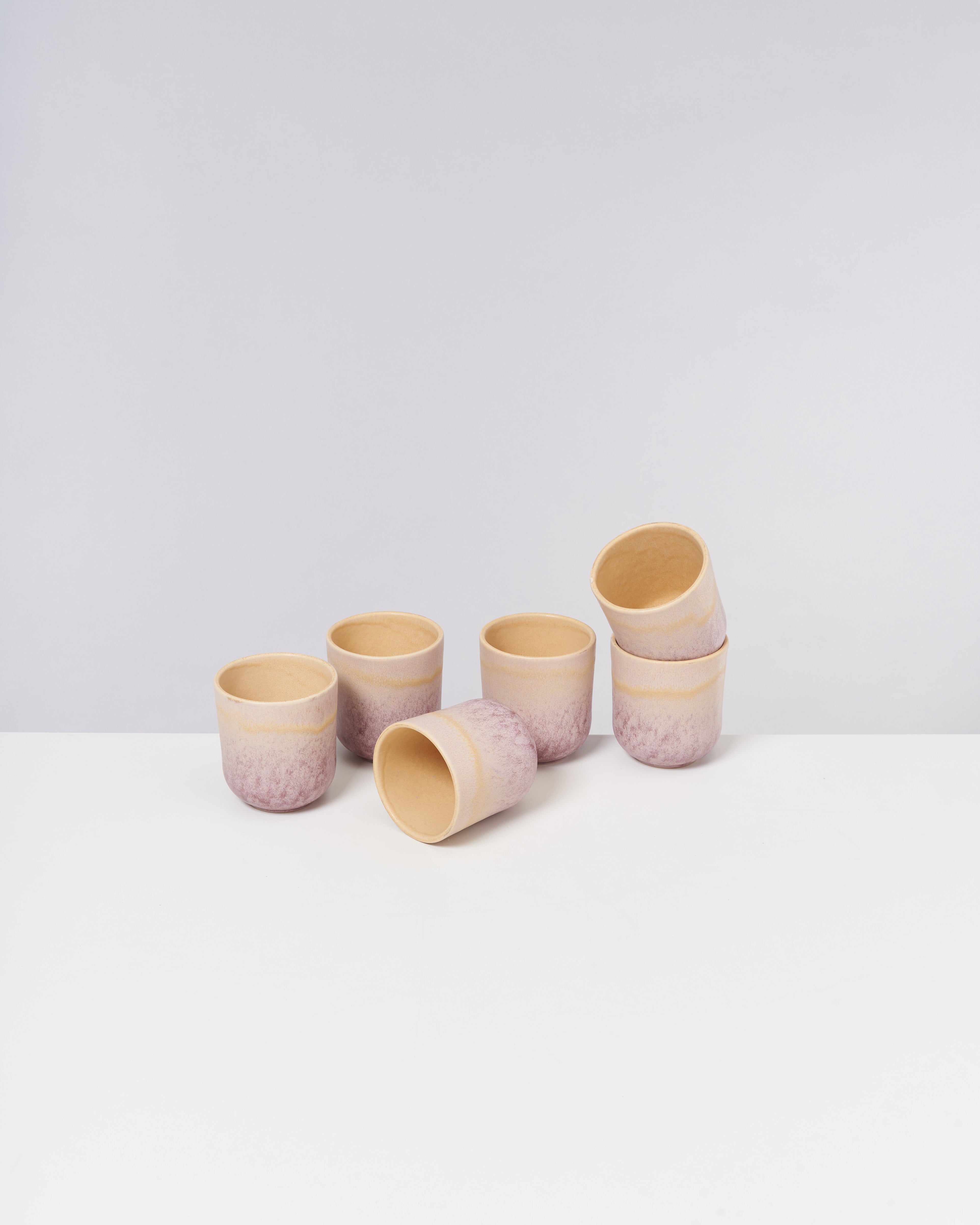 Sechs pastellfarbene Keramikbecher stehen auf einem weißen Tisch vor einem grauen Hintergrund.