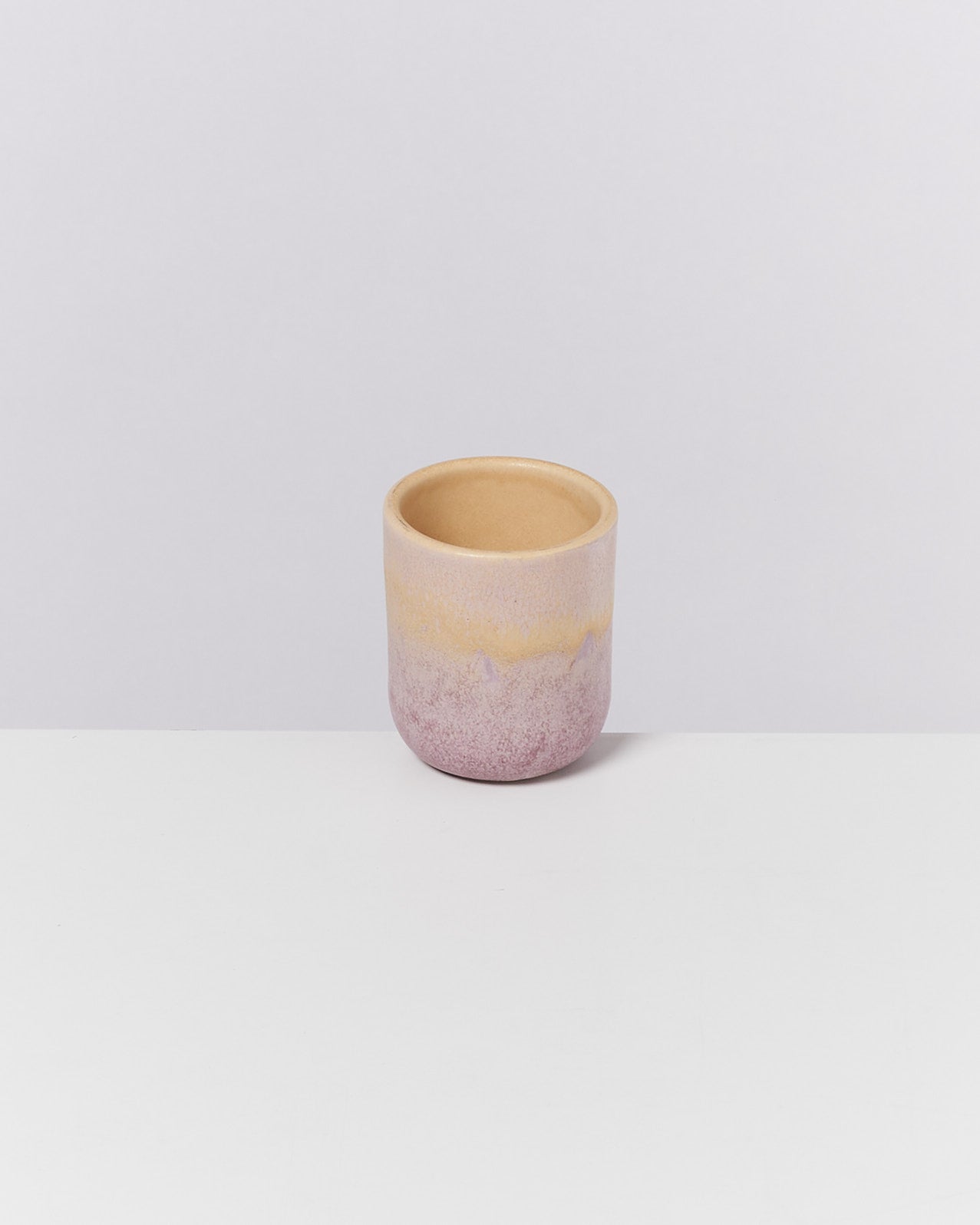 Kleine, bunte Keramiktasse mit rosa, gelber und lila Glasur auf einem weißen Untergrund vor grauem Hintergrund.