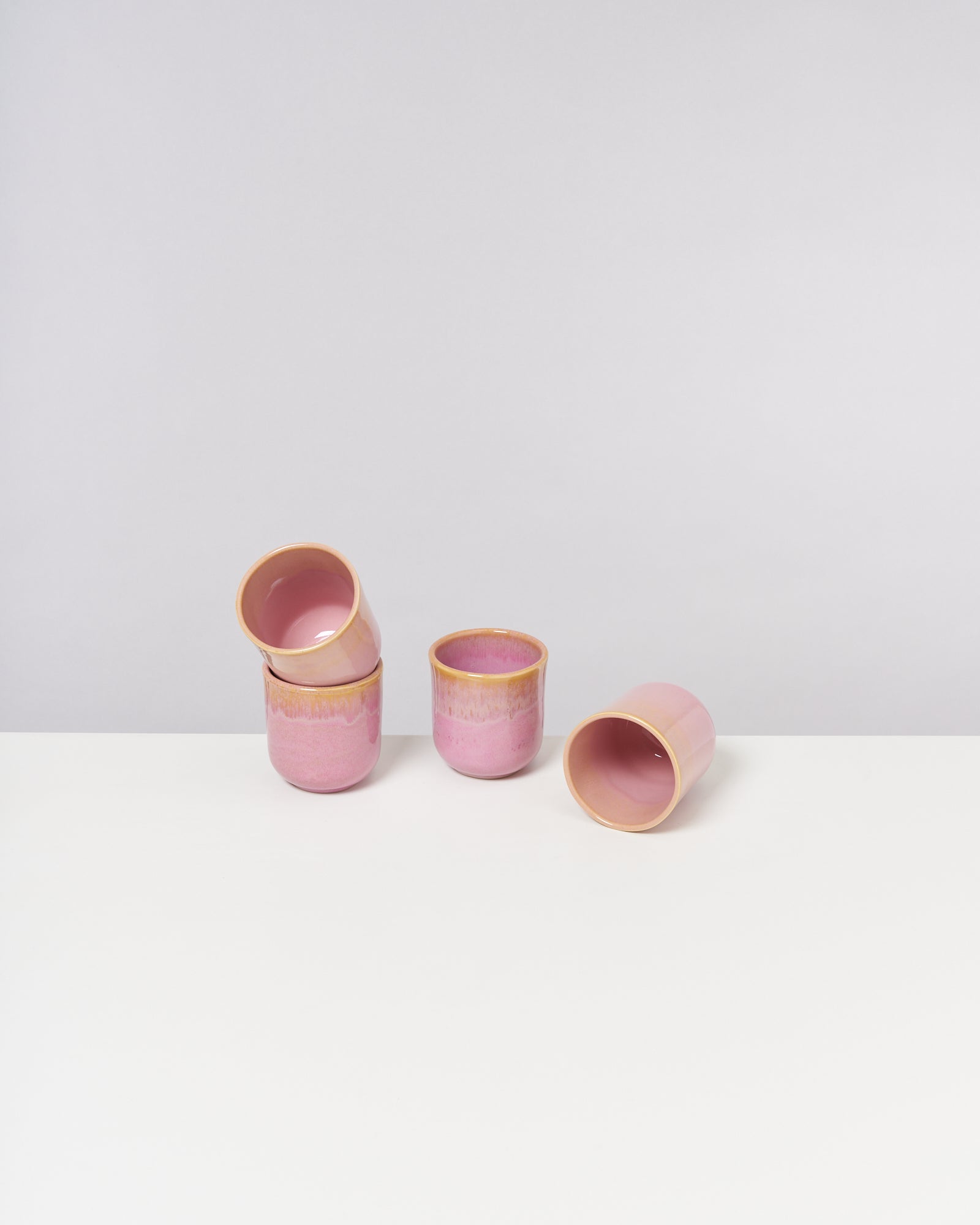 Vier rosafarbene Keramiktassen mit gelbem Rand stehen teilweise umgekippt auf einem weißen Tisch.