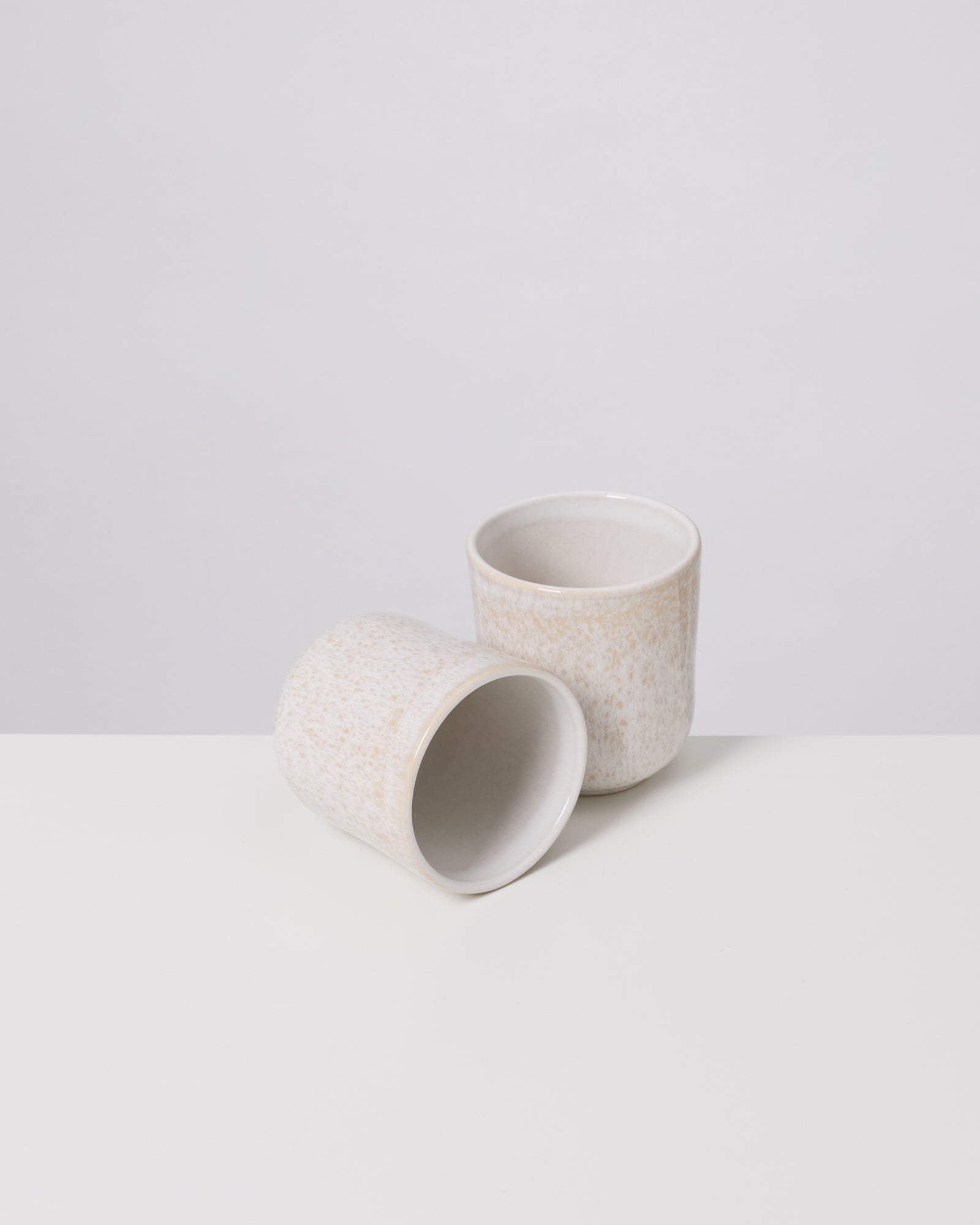 Zwei beige Keramiktassen, eine stehend und eine liegend, auf einer weißen Oberfläche vor einem grauen Hintergrund.