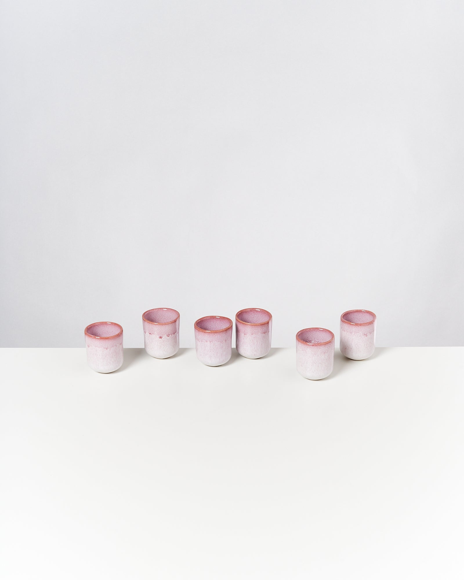 Sechs rosafarbene Keramikbecher stehen in einer Reihe auf einem weißen Tisch vor einem neutralen Hintergrund.